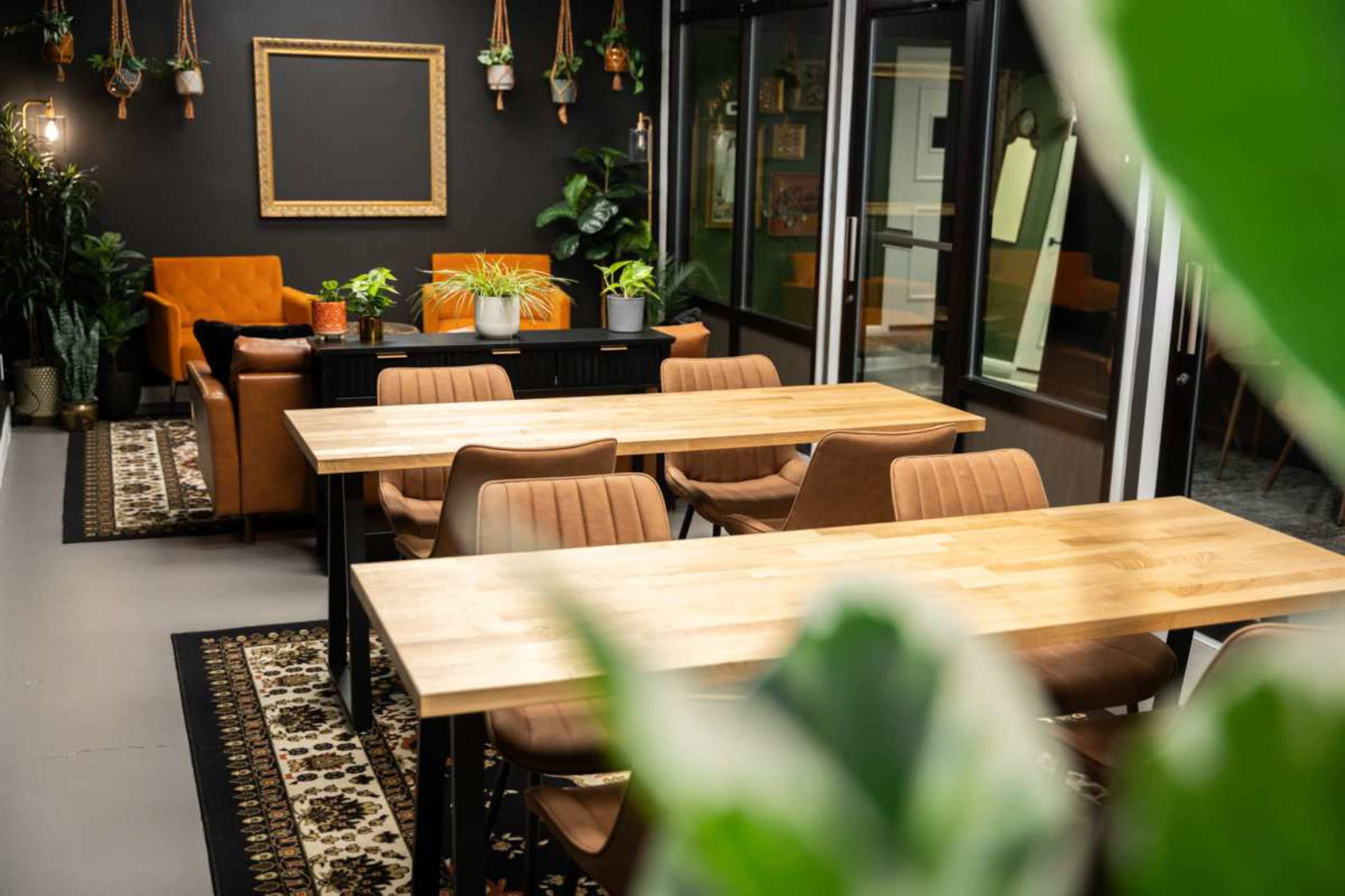 The image shows a modern, open space with wooden tables, brown chairs, and various plants, along with a comfortable seating area and a blank chalkboard on the wall.