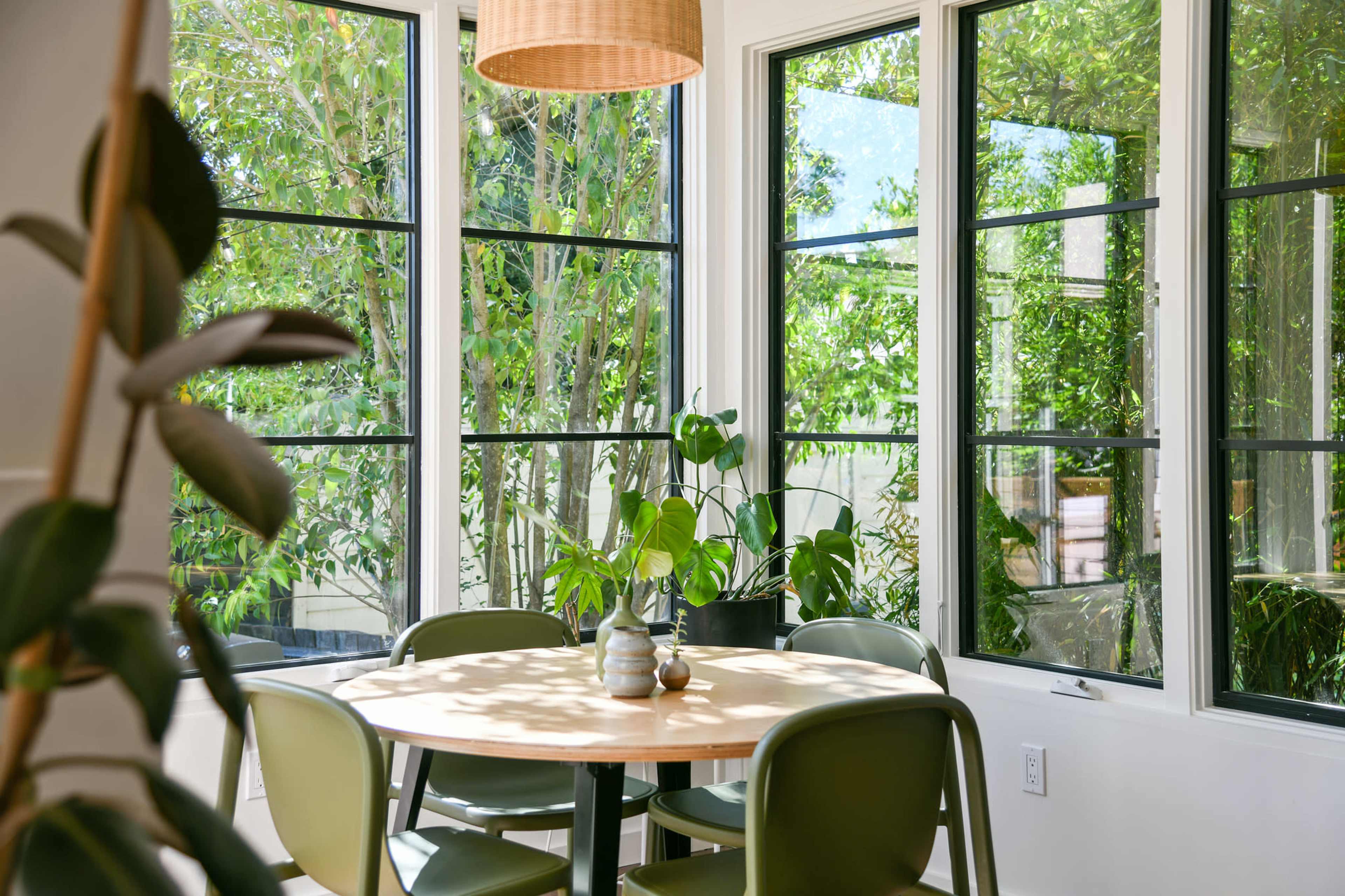 A round wooden table surrounded by green chairs is set in a sunlit room with large windows showcasing various indoor plants.