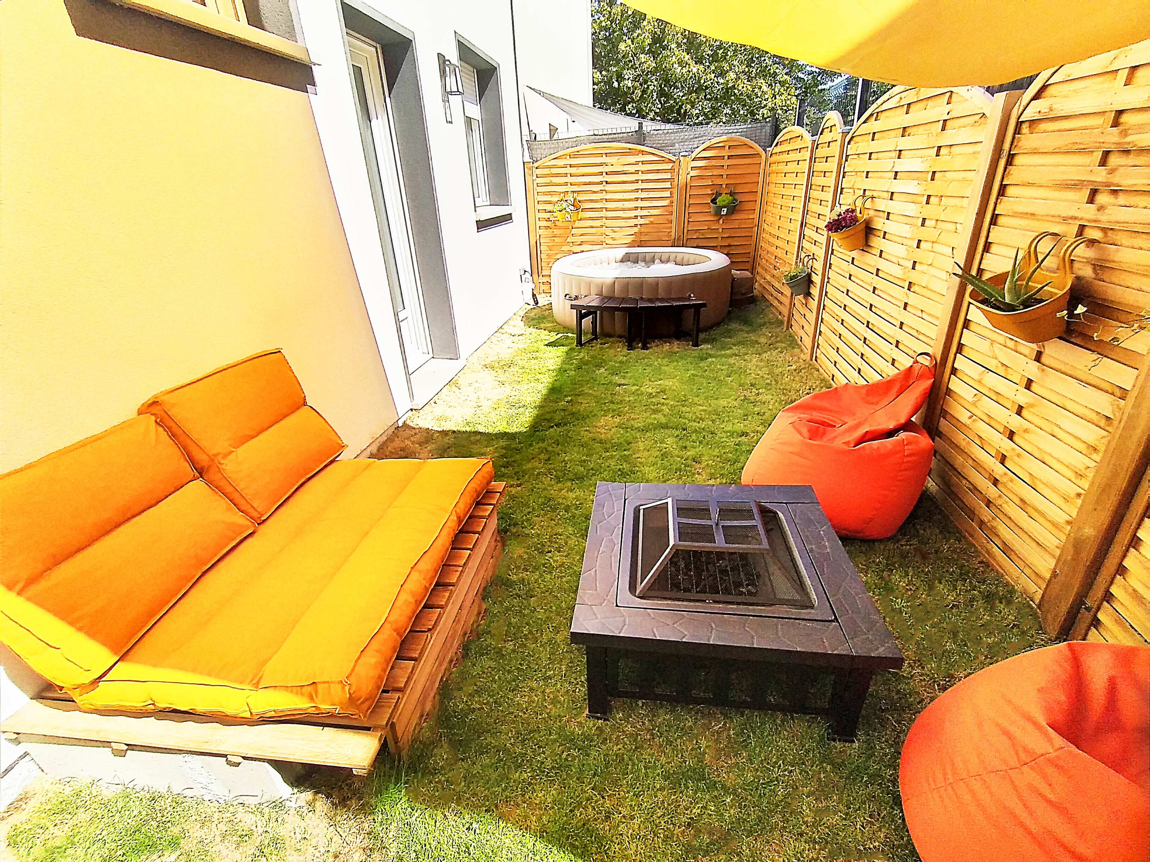 A small outdoor space with orange cushioned seating, a fire pit table, and scattered bean bags, surrounded by wooden fences and grass.