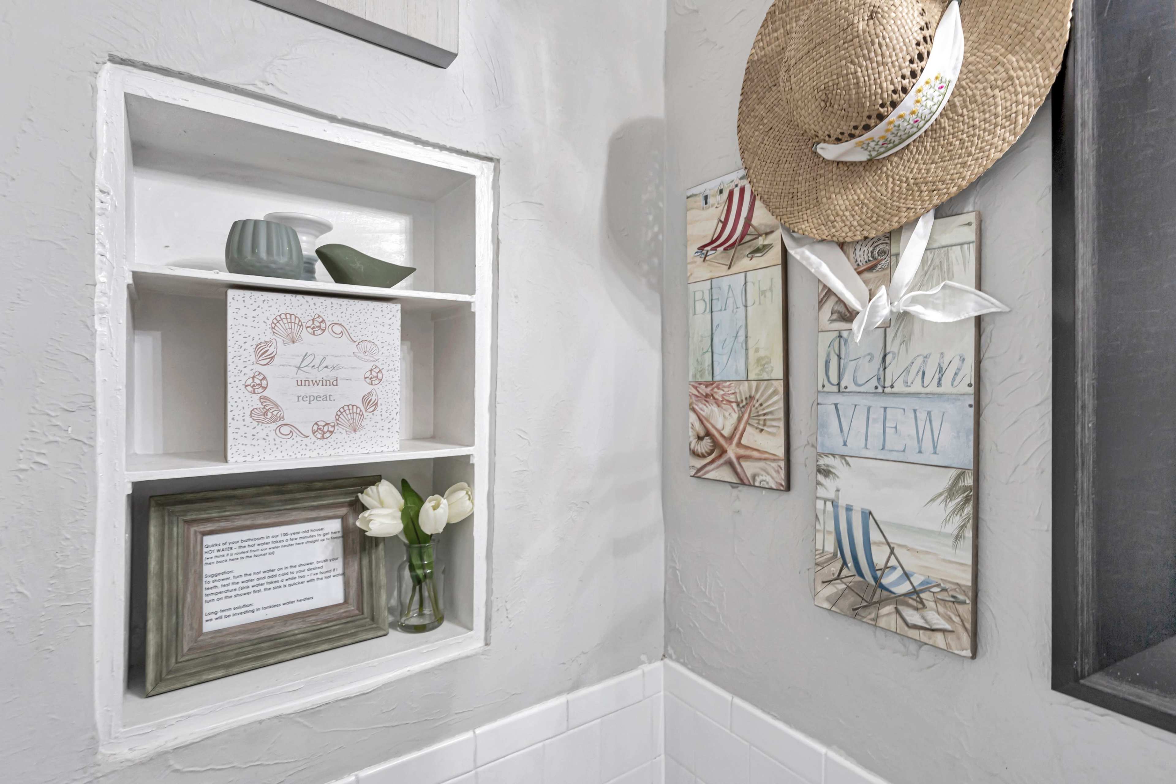 A bathroom corner featuring a wall shelf with decorative items, a framed message, and two beach-themed wall plaques.
