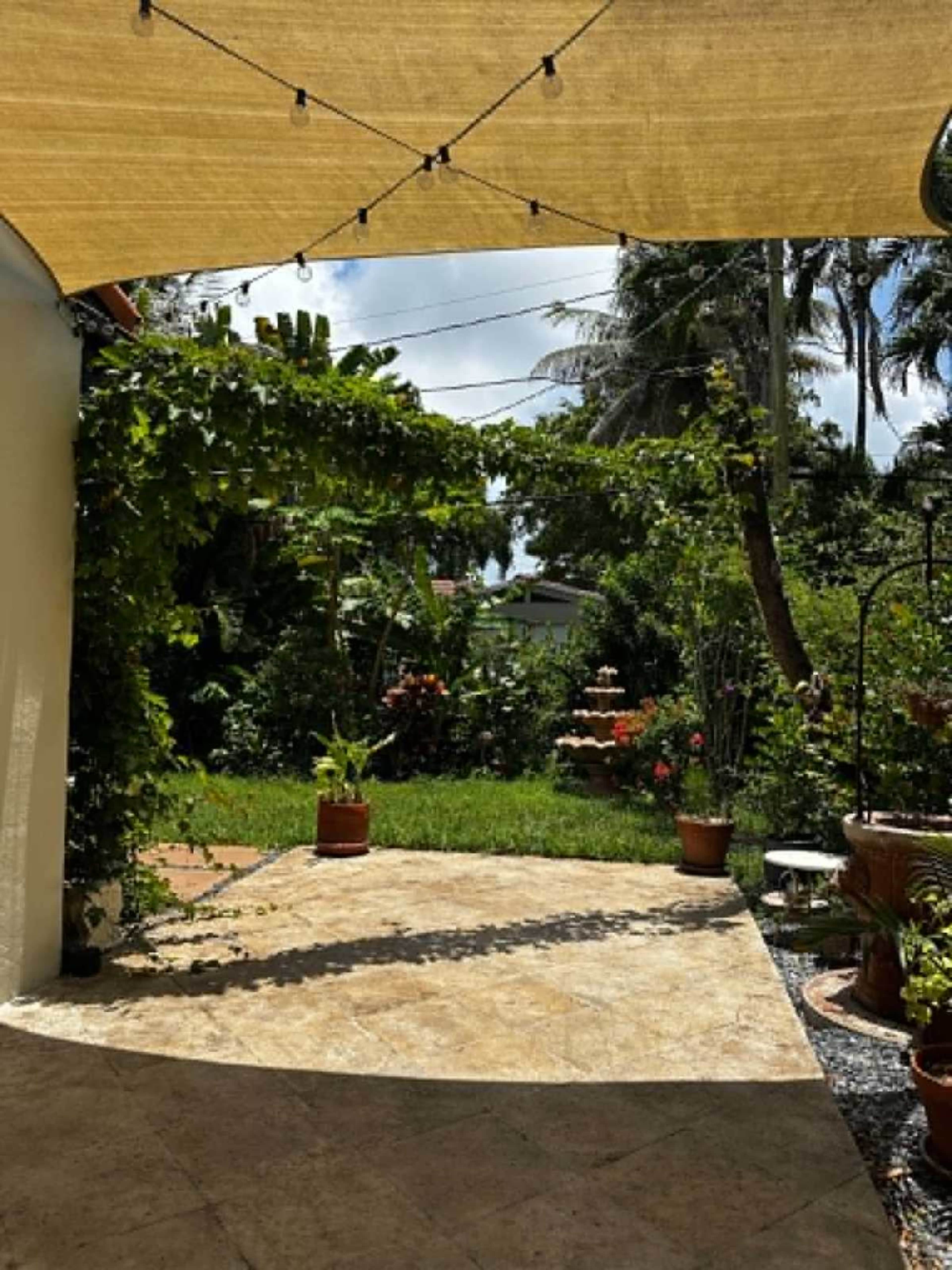 A shaded outdoor area features a stone patio with potted plants and a lush garden in the background.