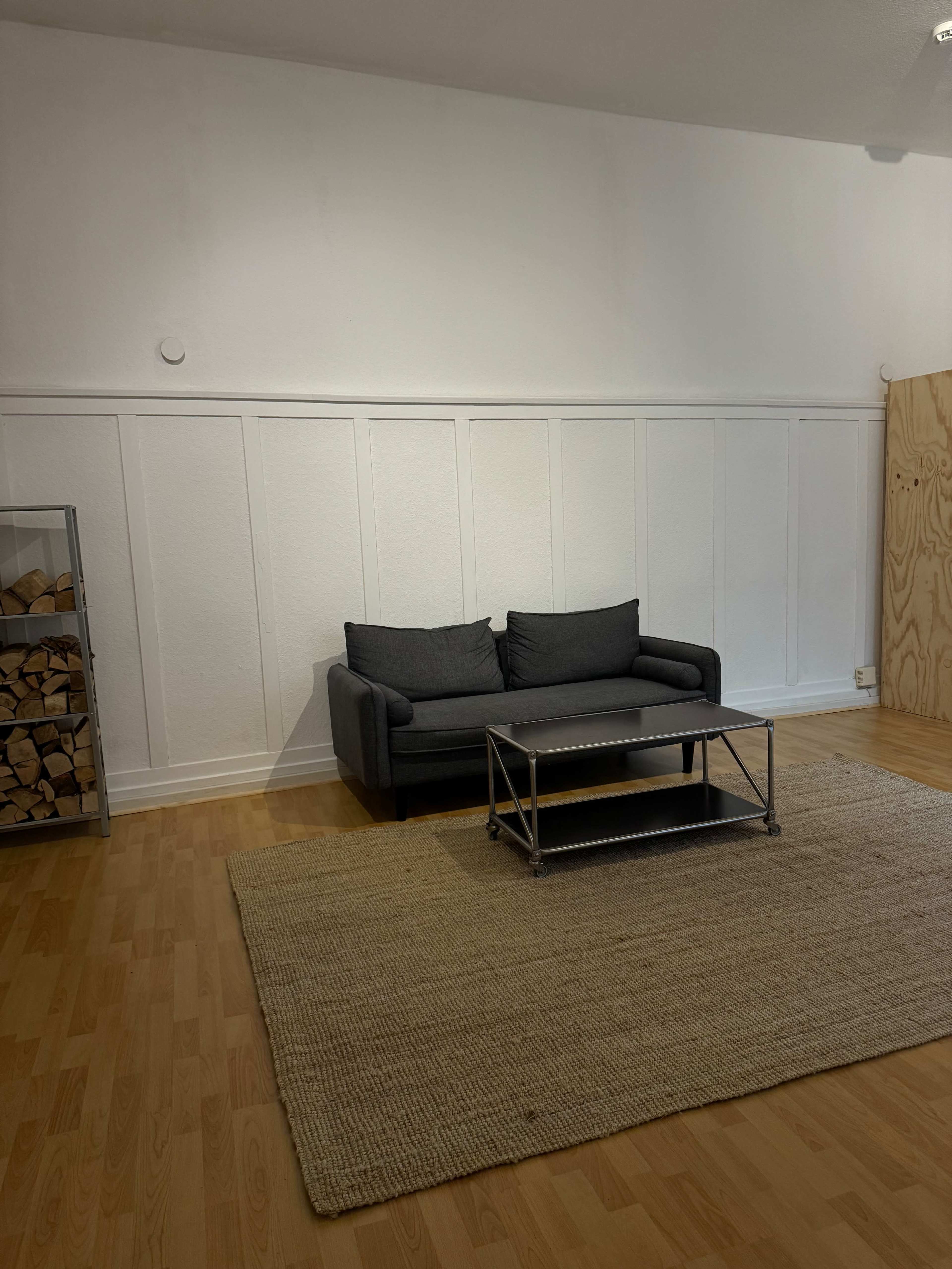 A minimalist living space features a gray sofa, a coffee table, and a large woven rug on a wooden floor, with wooden logs displayed in a nearby glass cabinet.