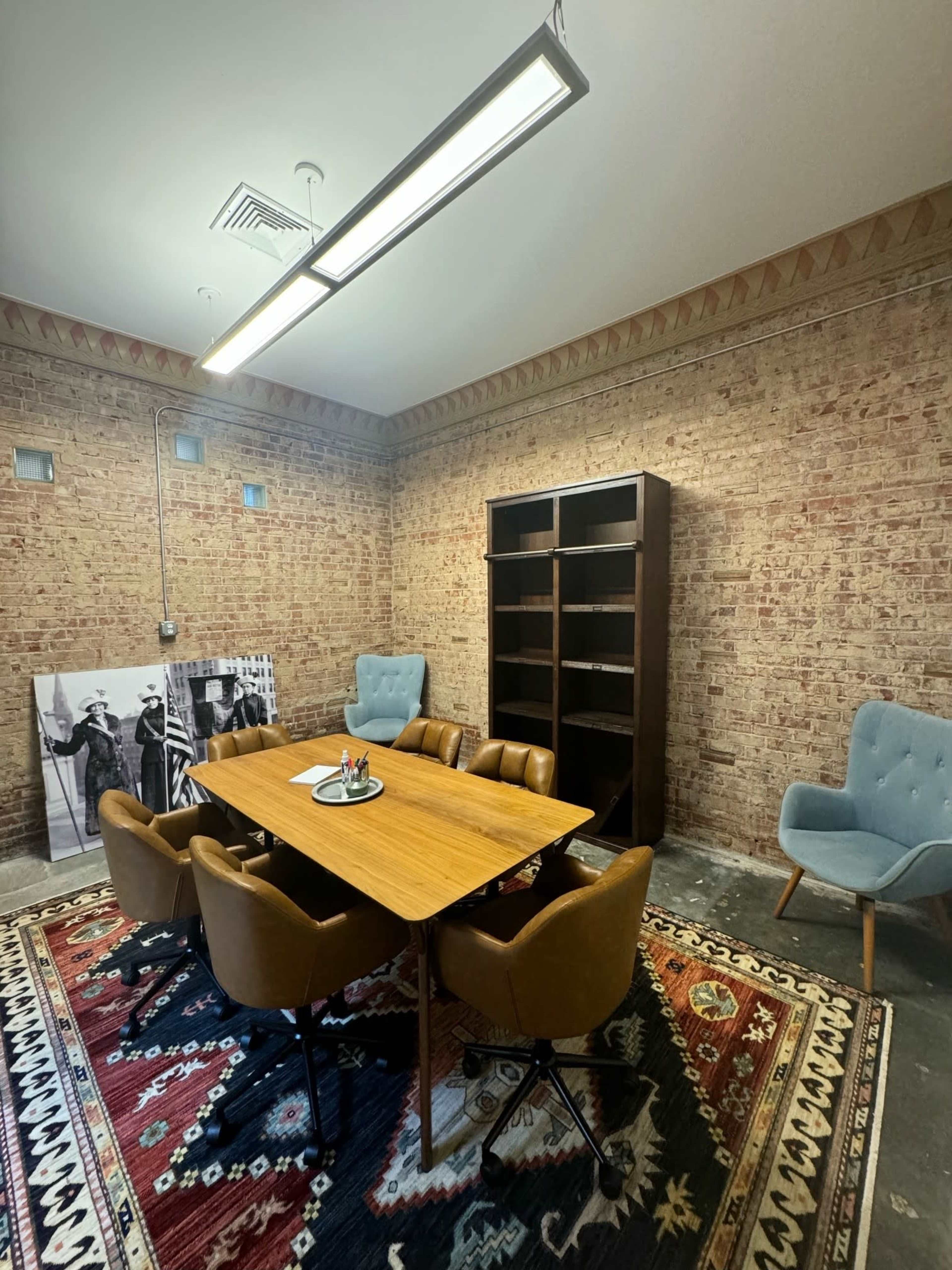 A meeting room features a large wooden table surrounded by brown chairs, with a patterned rug on the floor and a bookshelf against a brick wall.