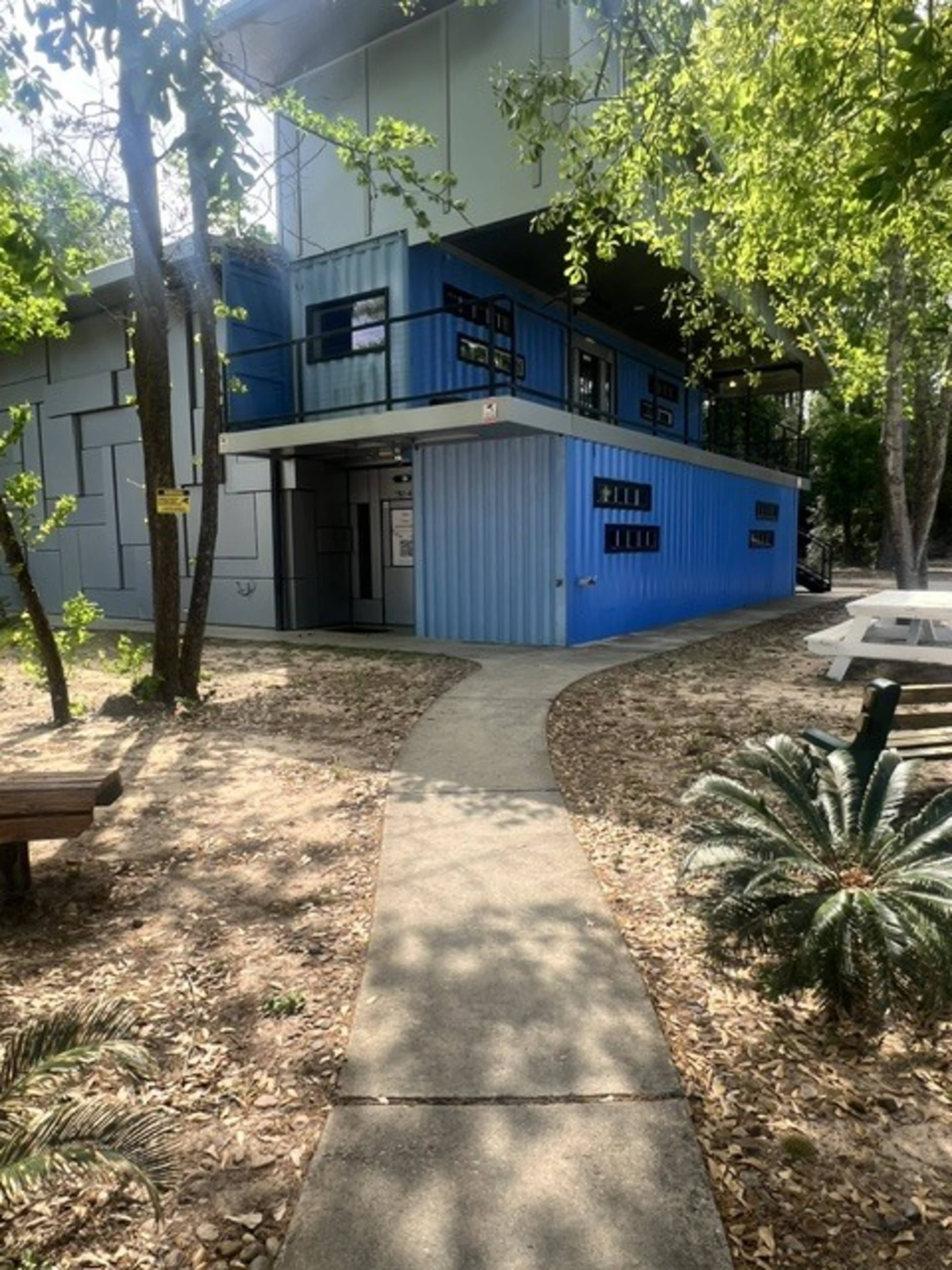 A walkable pathway leads to a modern building constructed with blue shipping containers, surrounded by trees and outdoor seating areas.
