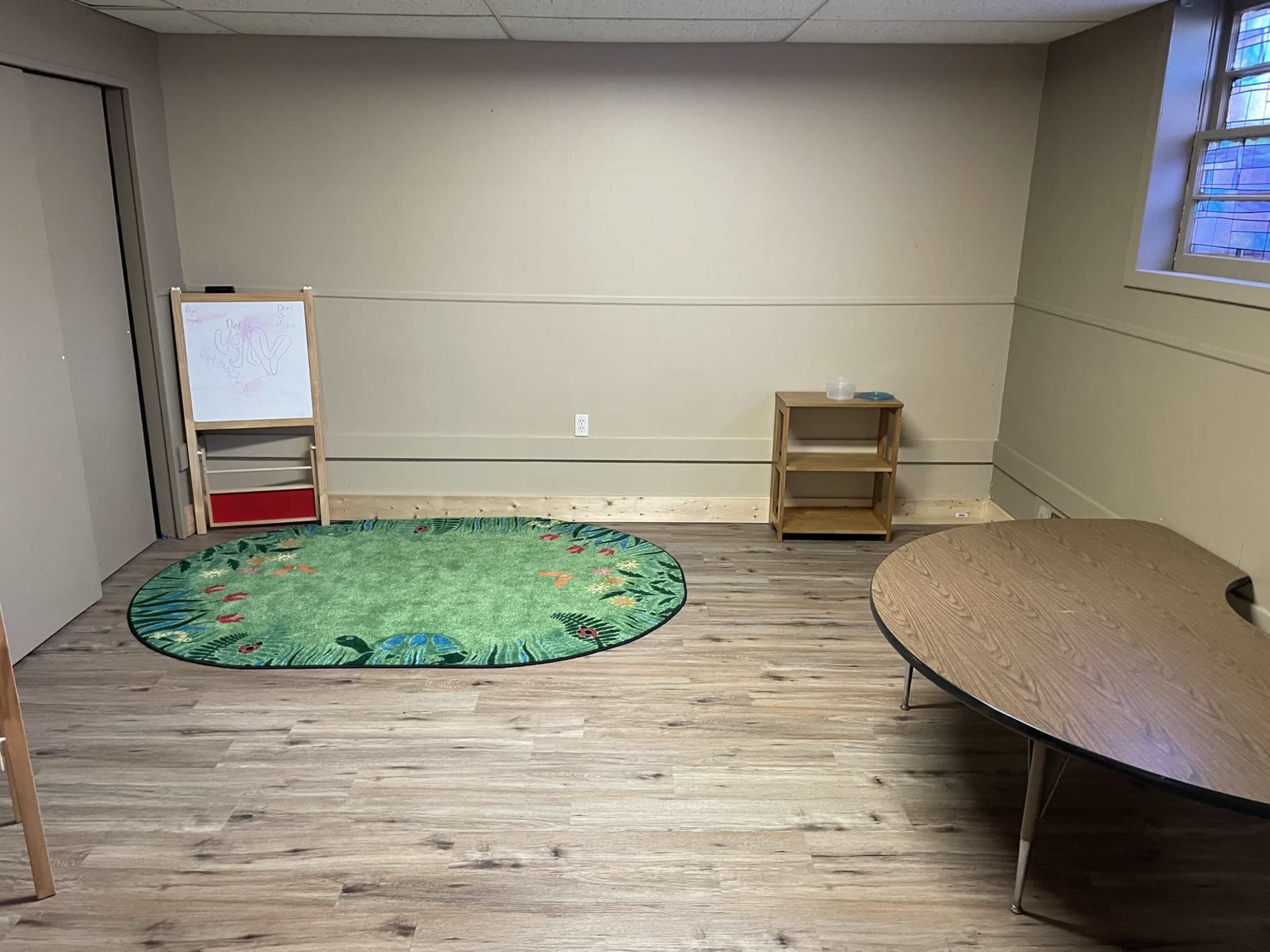 The room features a green circular rug with a nature design, a wooden table, a bookshelf, and a whiteboard on an easel.