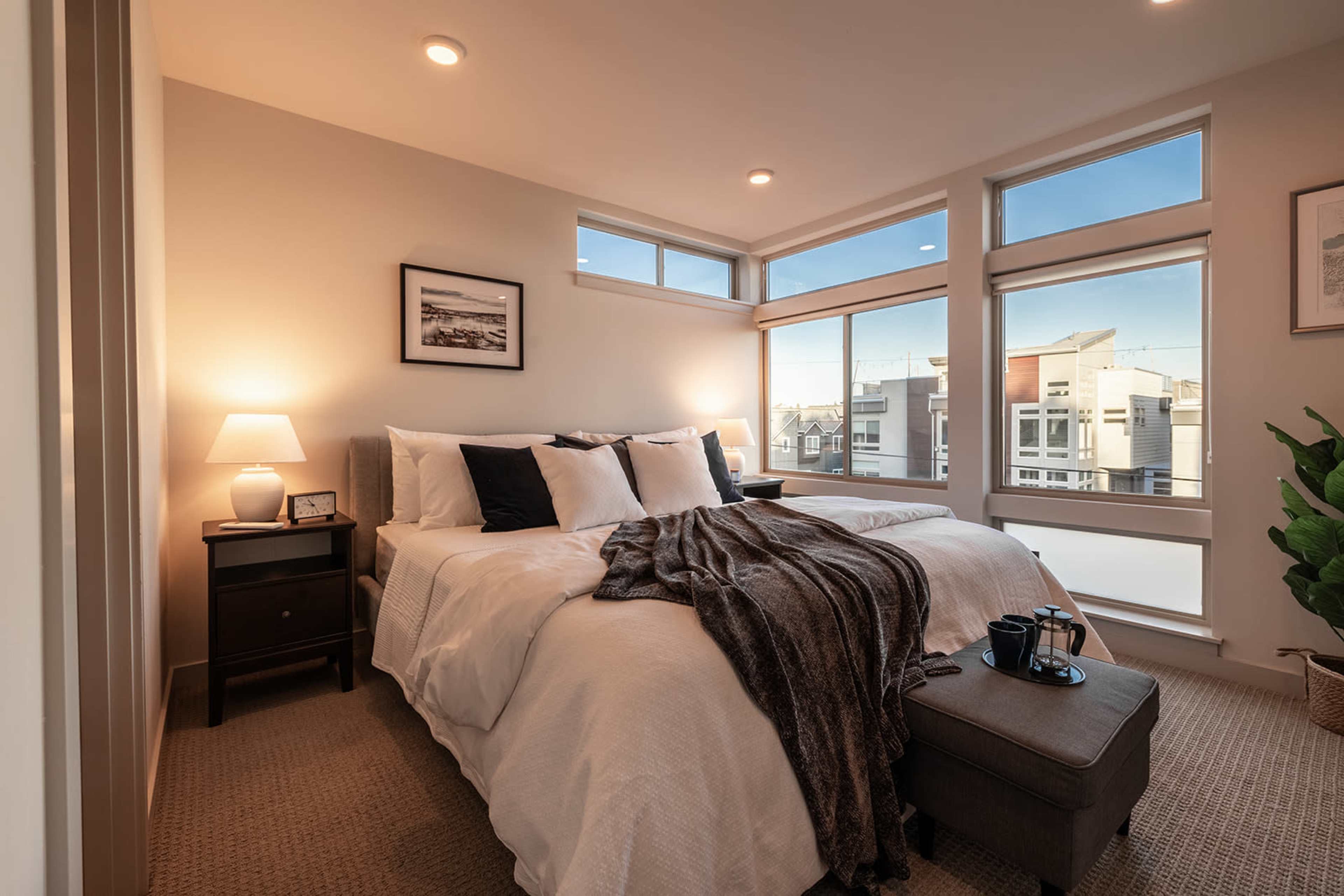 A well-lit bedroom features a large bed with white linens, a cozy throw blanket, a bedside table with a lamp, and windows framing a view of the outside.