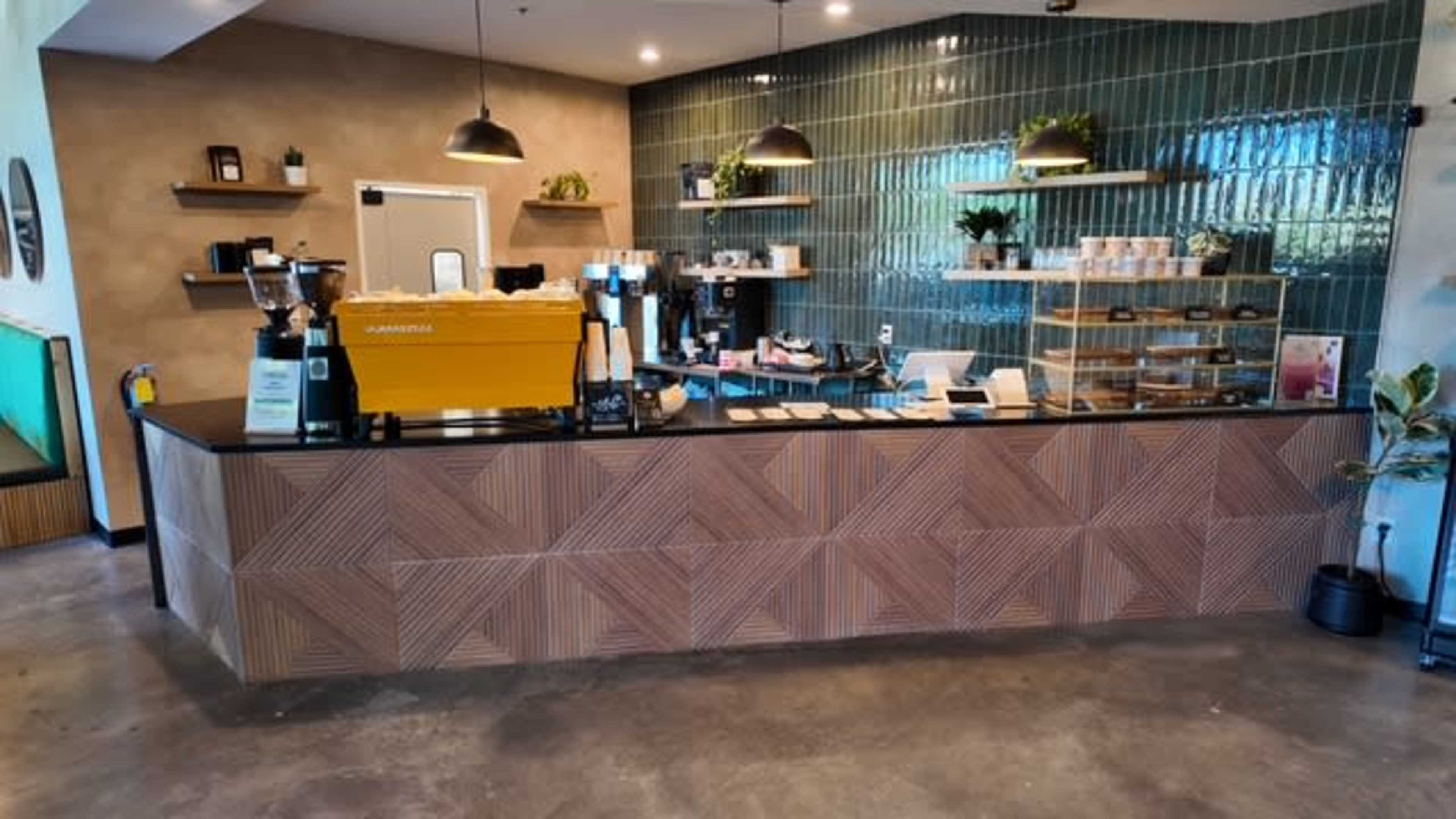 A modern coffee shop interior features a sleek counter with an espresso machine and a display of baked goods against a backdrop of green tile and wooden shelves.