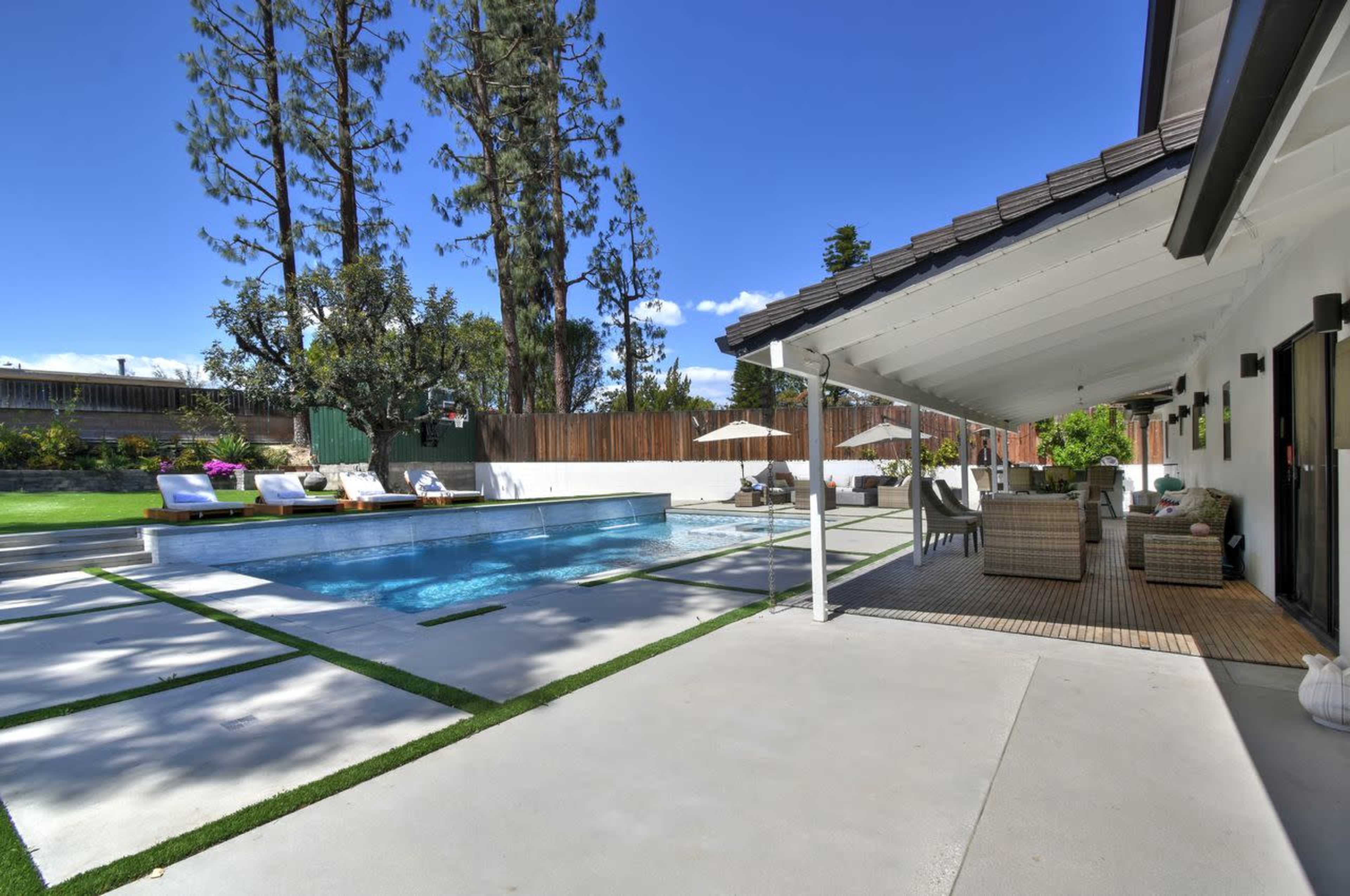 The image shows a backyard with a swimming pool, lounge chairs, and a covered seating area, surrounded by trees and a wooden fence.