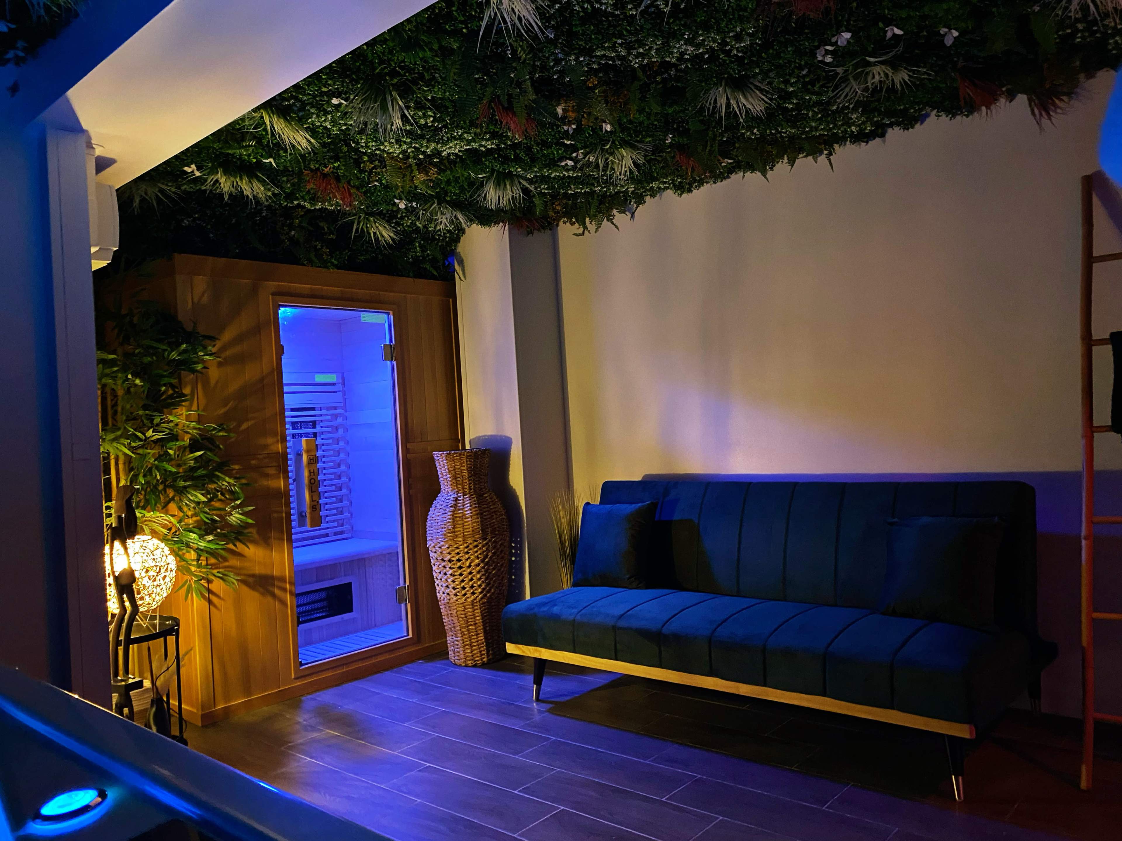 The scene features a modern room with a green wall of plants, a blue-lit sauna, a blue sofa, and decorative vases.