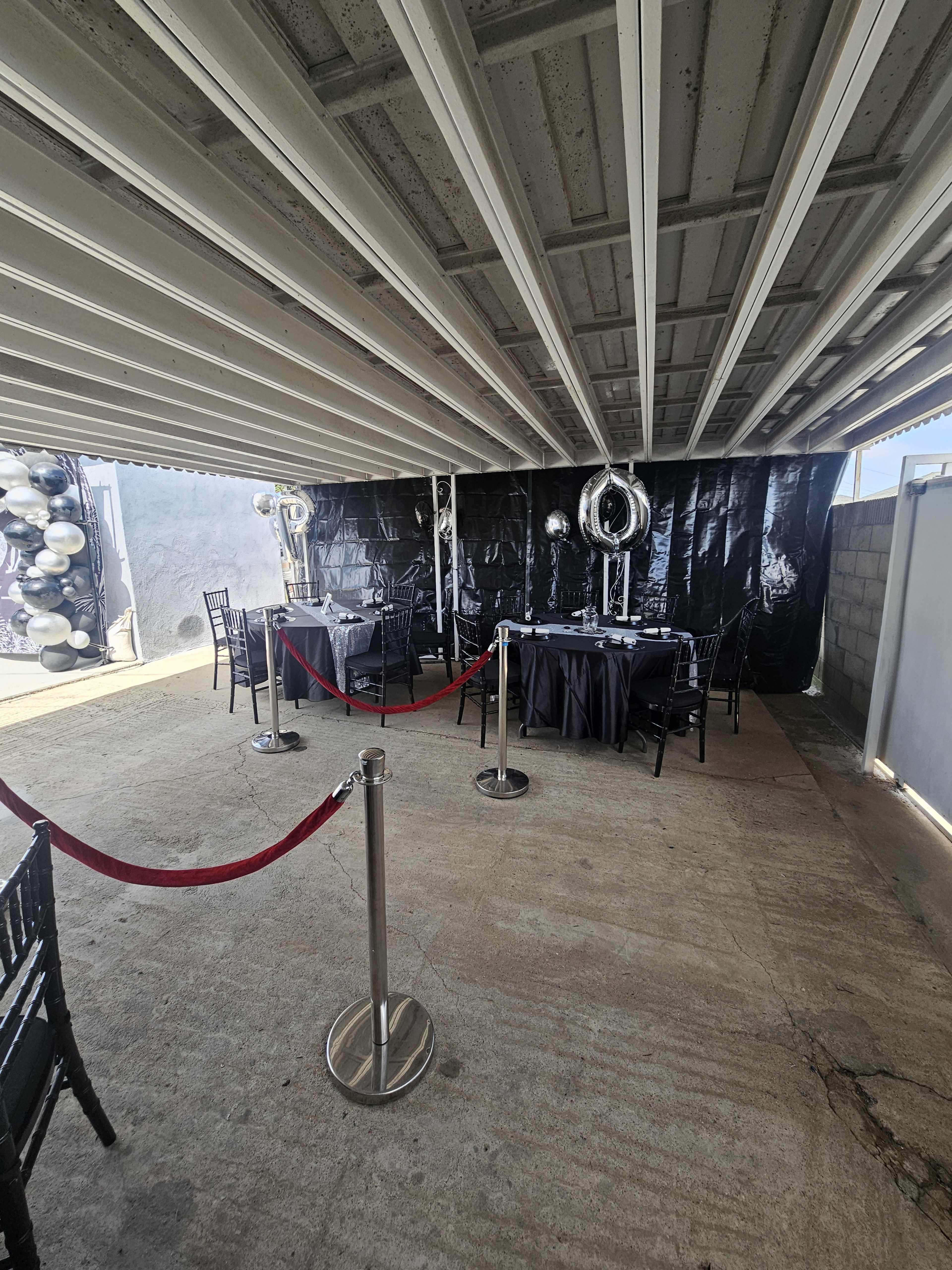 A tented area features tables covered with black cloth, surrounded by black chairs and silver balloons, with a red rope barrier at the entrance.