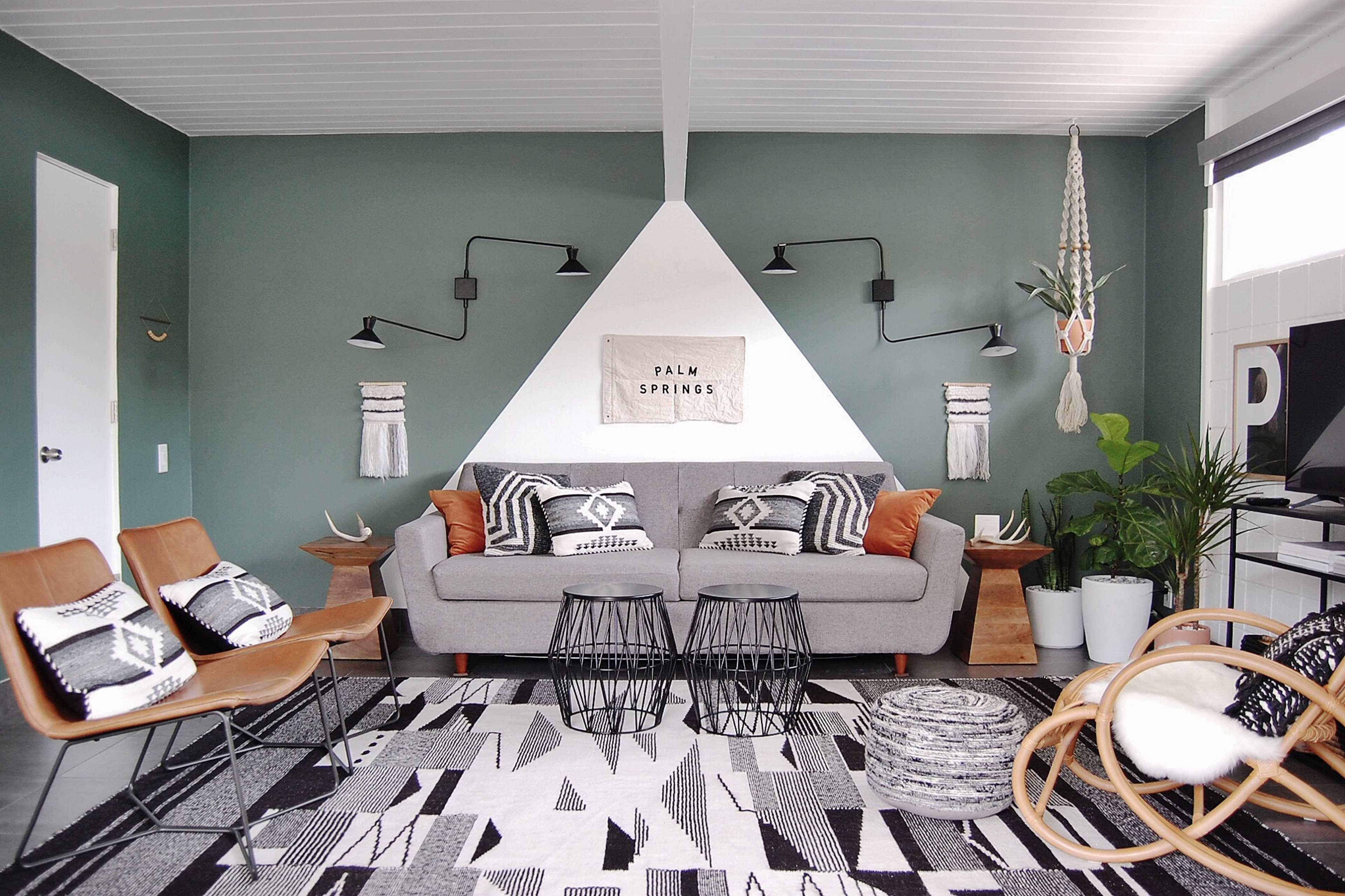 A modern living room features a gray sofa with patterned pillows, two geometric side tables, and an area rug with black and white designs, set against a green accent wall.