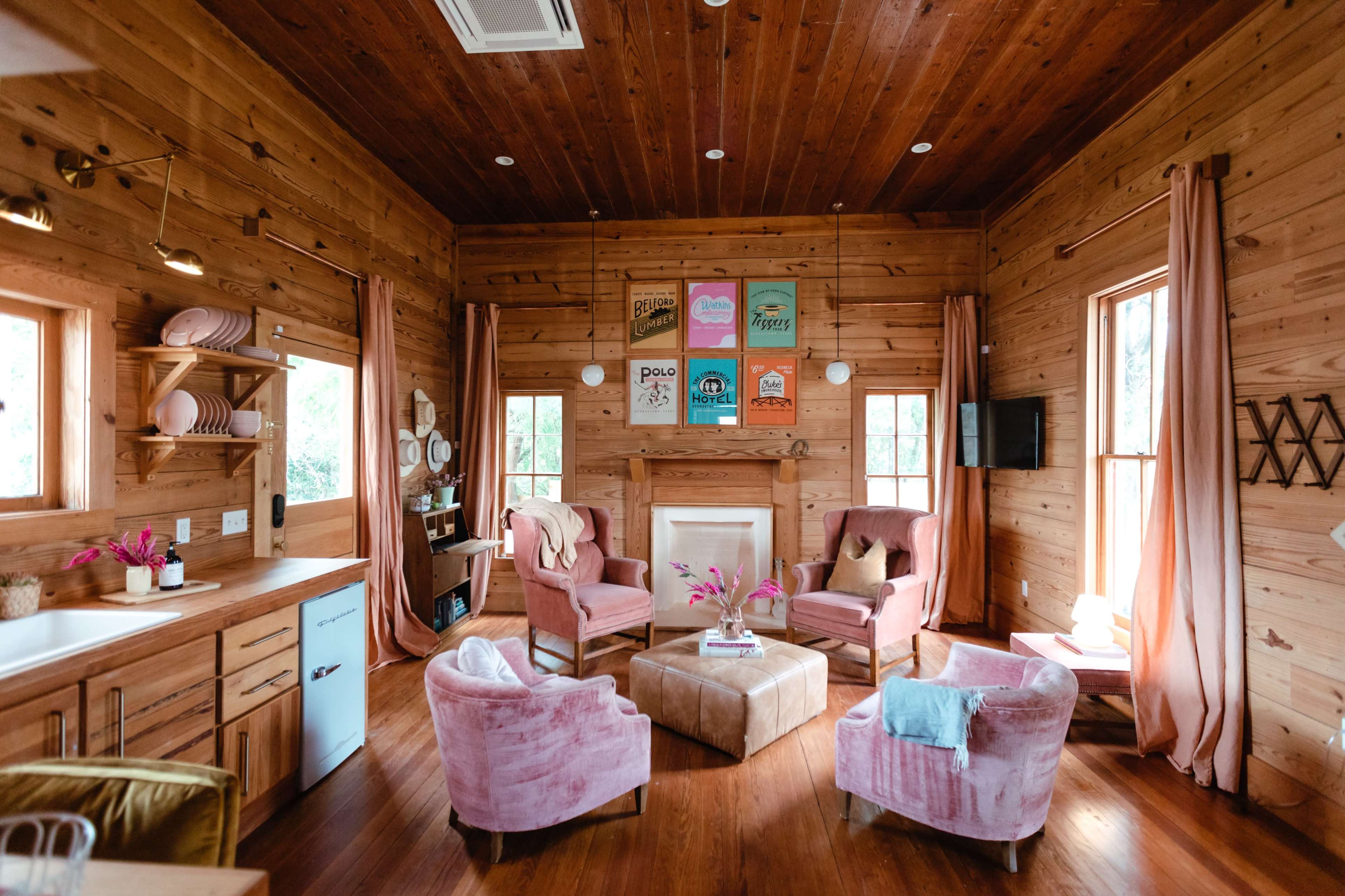 A cozy wooden cabin living room with pink upholstered chairs, a coffee table, and colorful artwork on the walls.