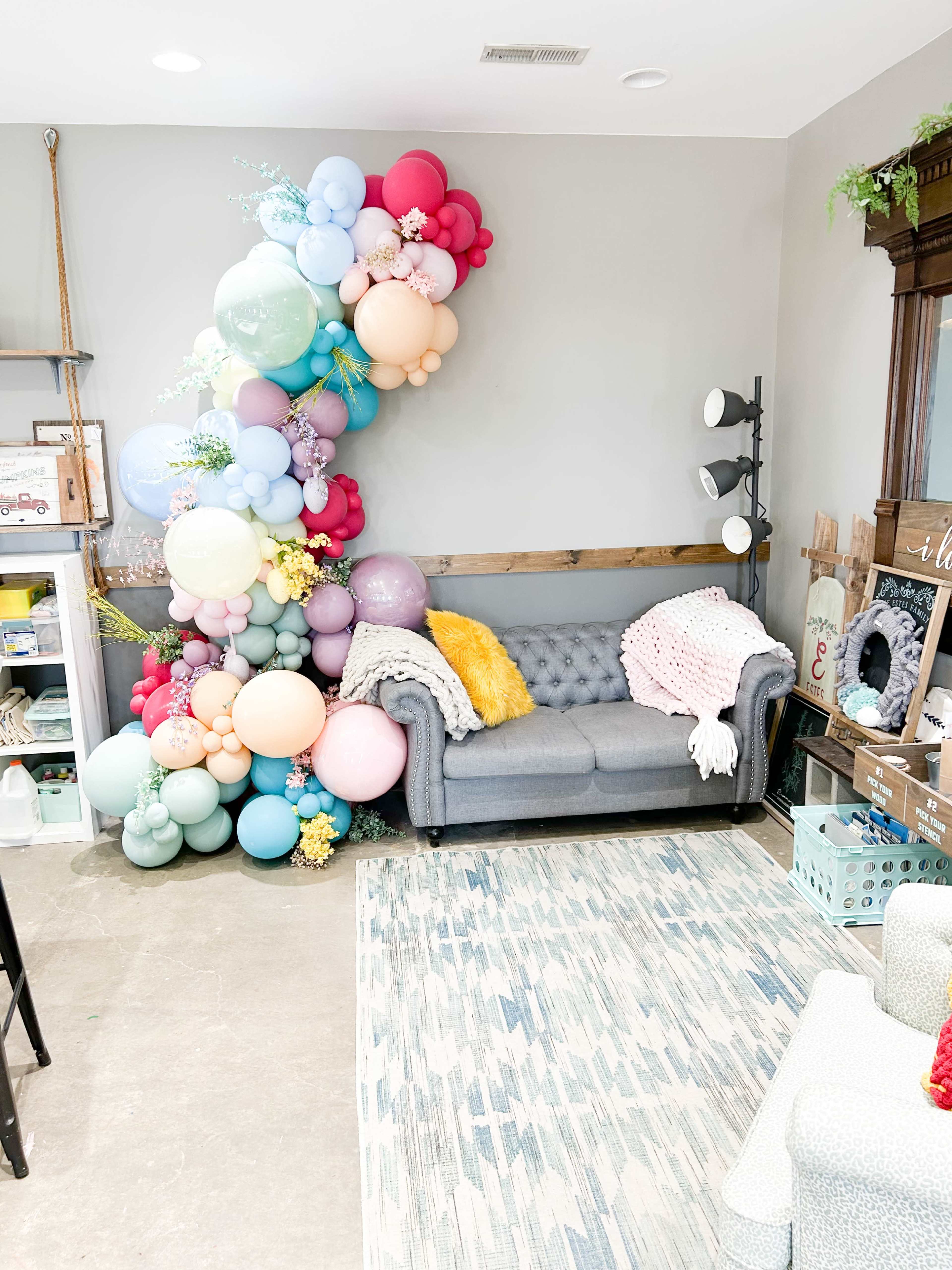A cozy living space featuring a gray tufted sofa adorned with a colorful arrangement of balloons in various sizes and colors along one wall, complemented by a patterned rug and a shelf with decorative items.