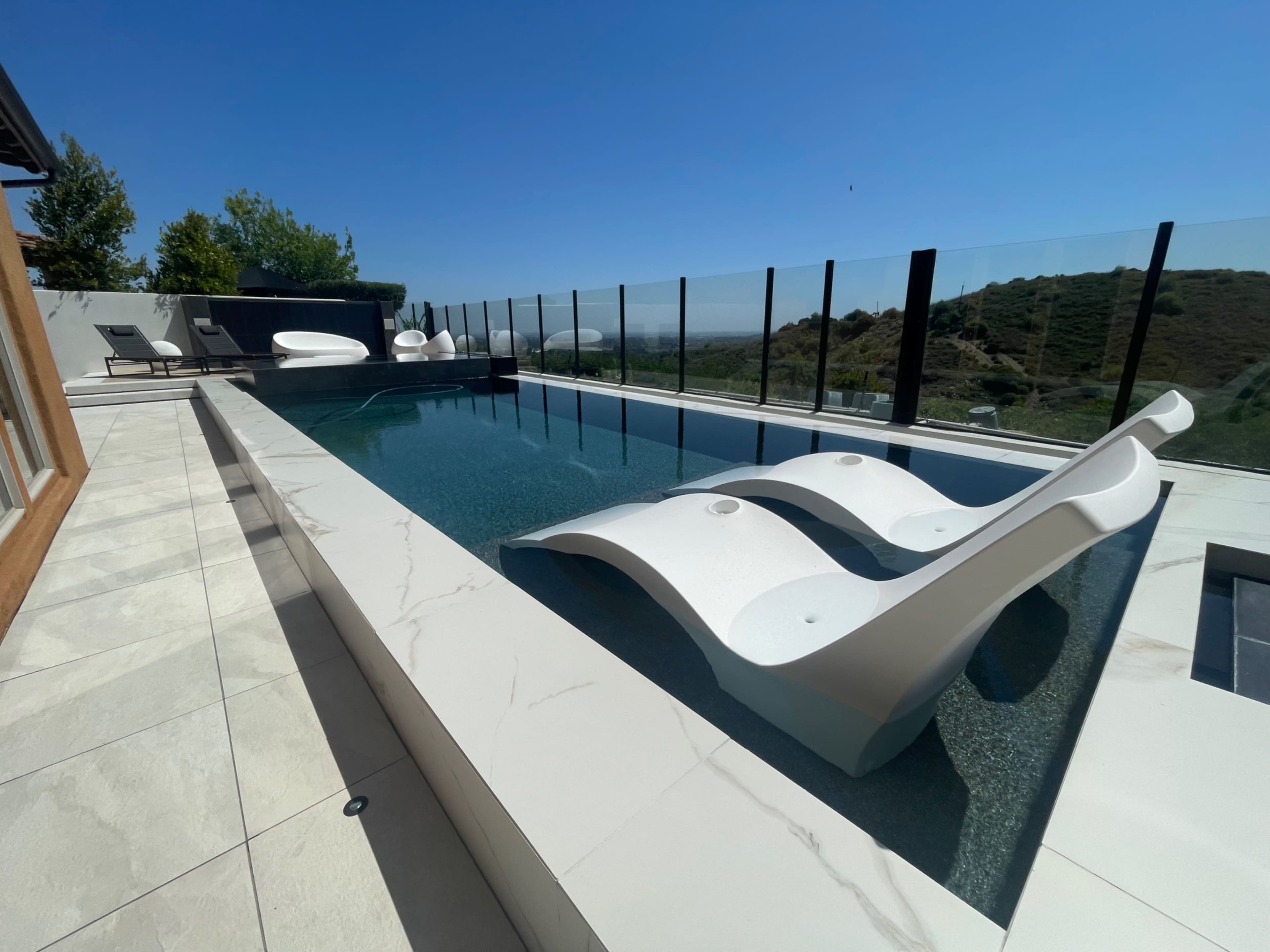 The image shows a modern infinity pool with white loungers and a clear glass railing overlooking a hillside.