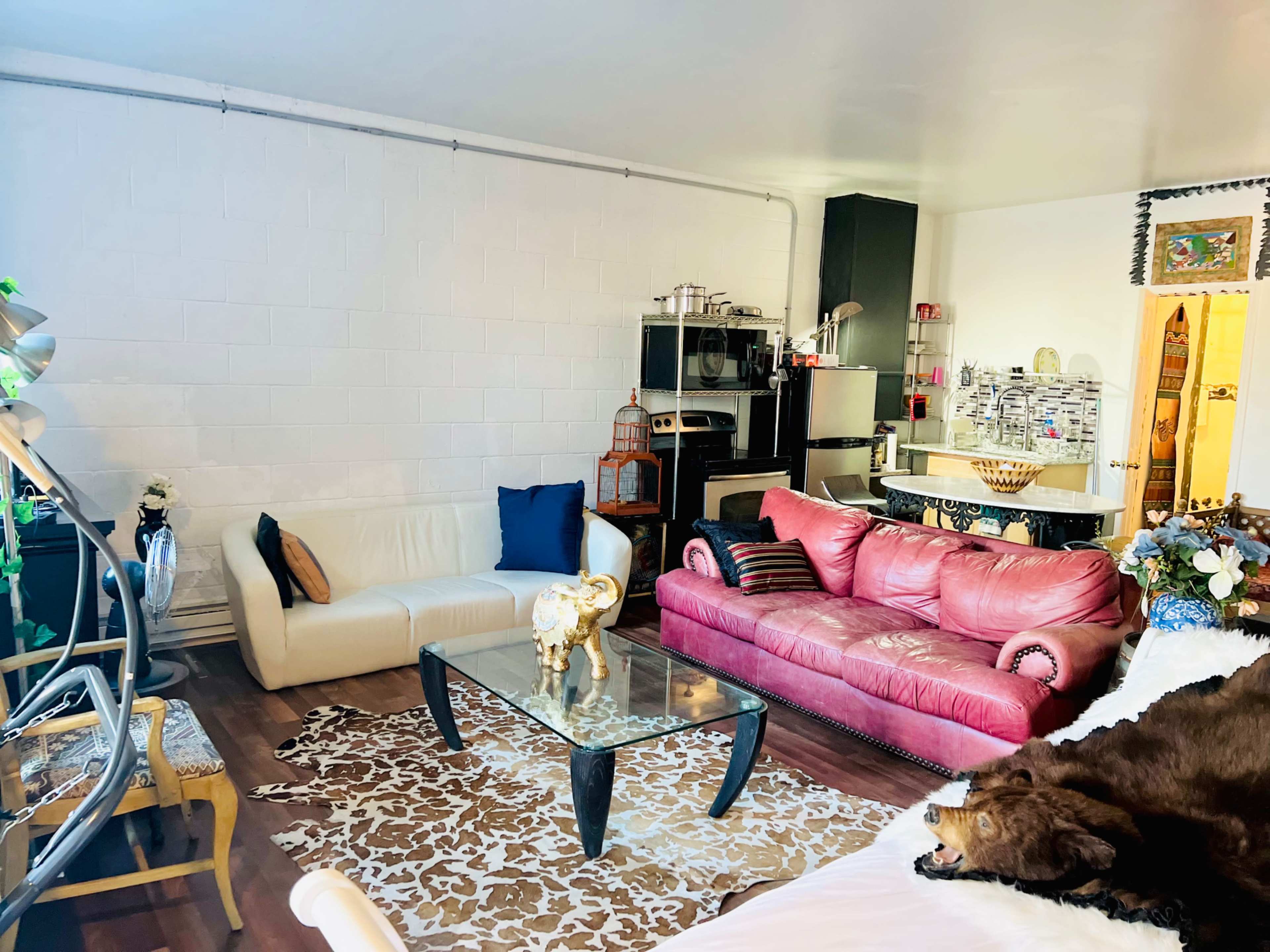 A cozy living room with a white couch, a pink leather sofa, a glass coffee table on a patterned rug, and a view of a kitchen area in the background.
