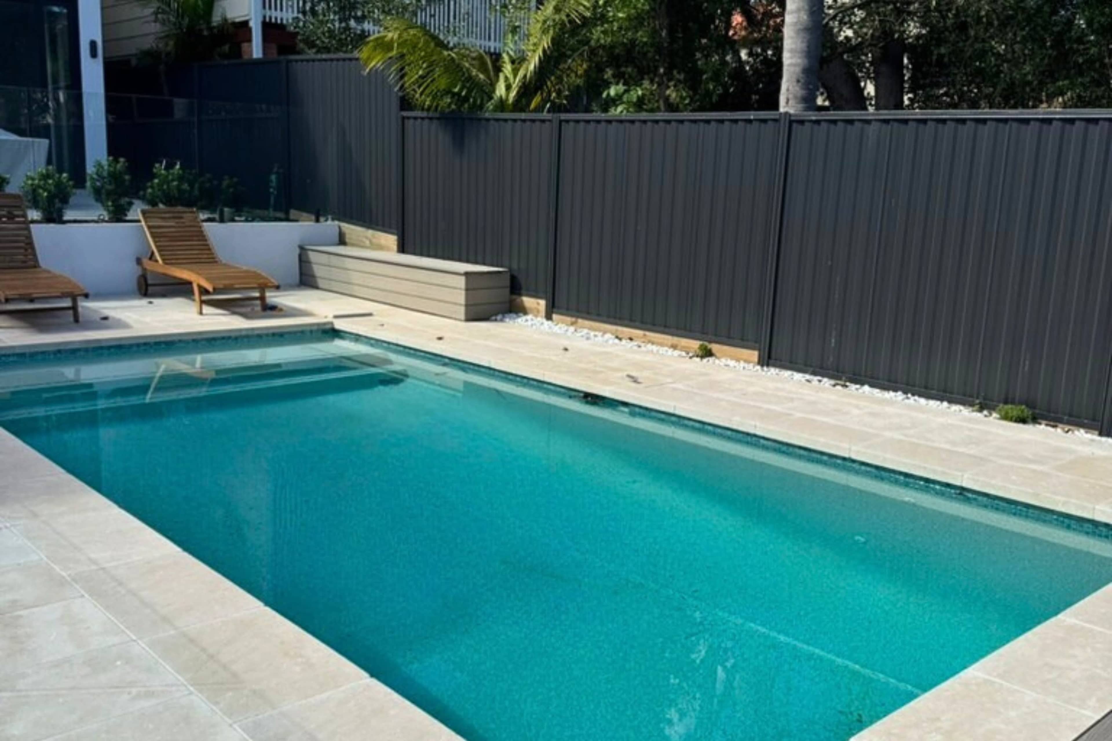 The image shows a modern swimming pool surrounded by light-colored patio tiles and two wooden lounge chairs, with a black fence in the background.