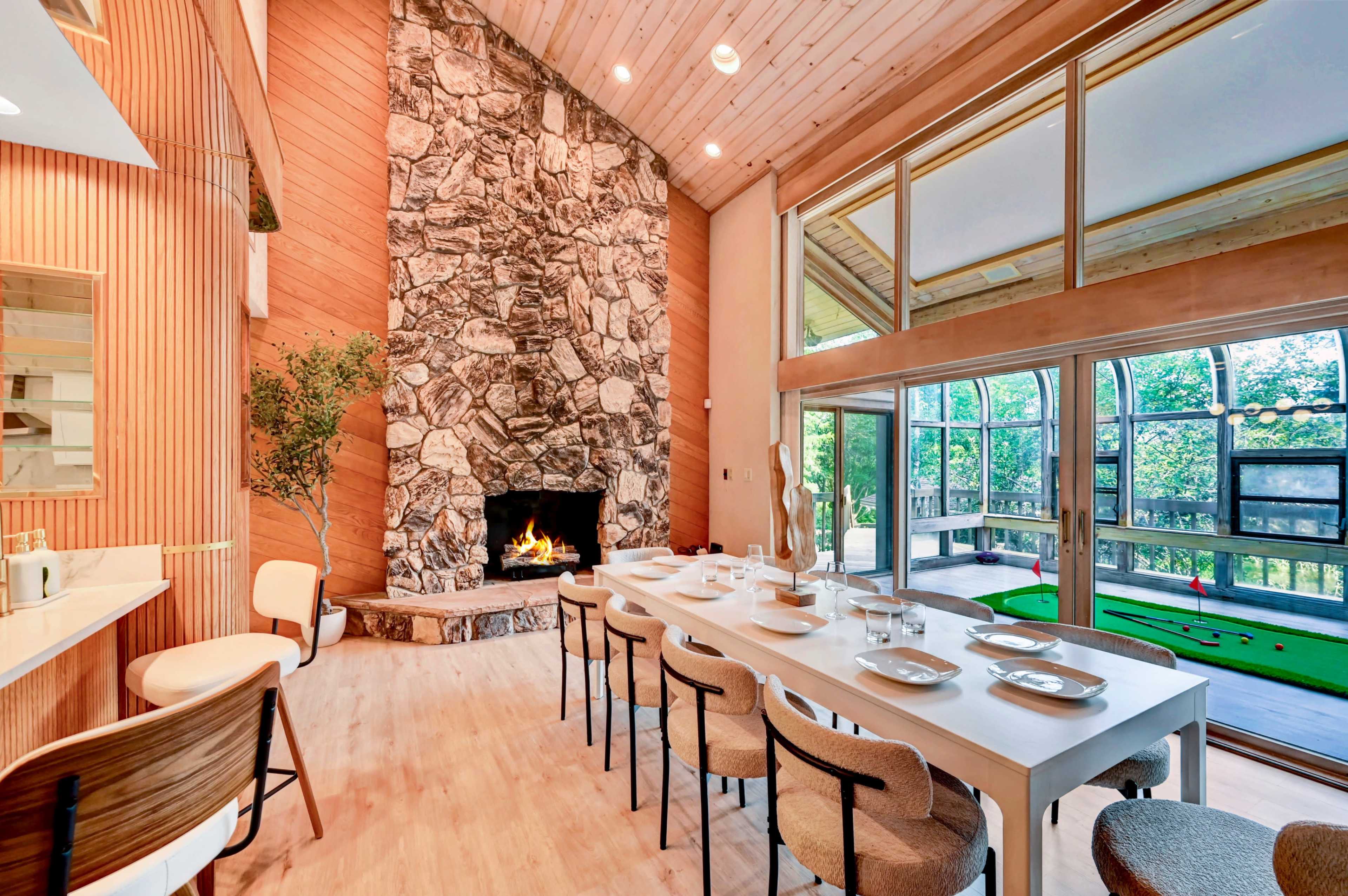 The image shows a modern dining area with a stone fireplace and large windows overlooking a lush outdoor space.