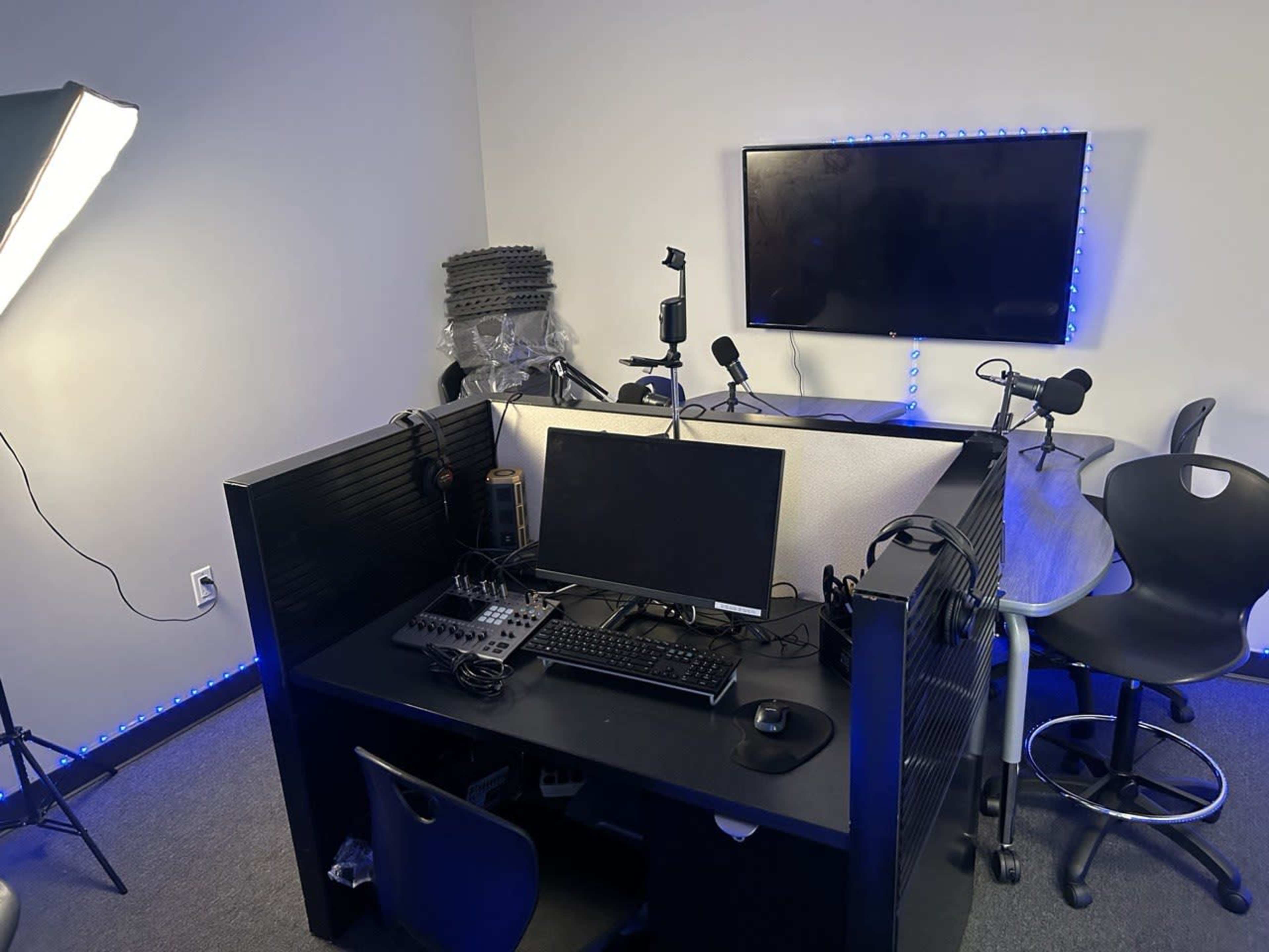 The image shows a small recording studio setup with a computer, audio mixing equipment, and microphones, surrounded by soundproof panels and illuminated by blue lights.