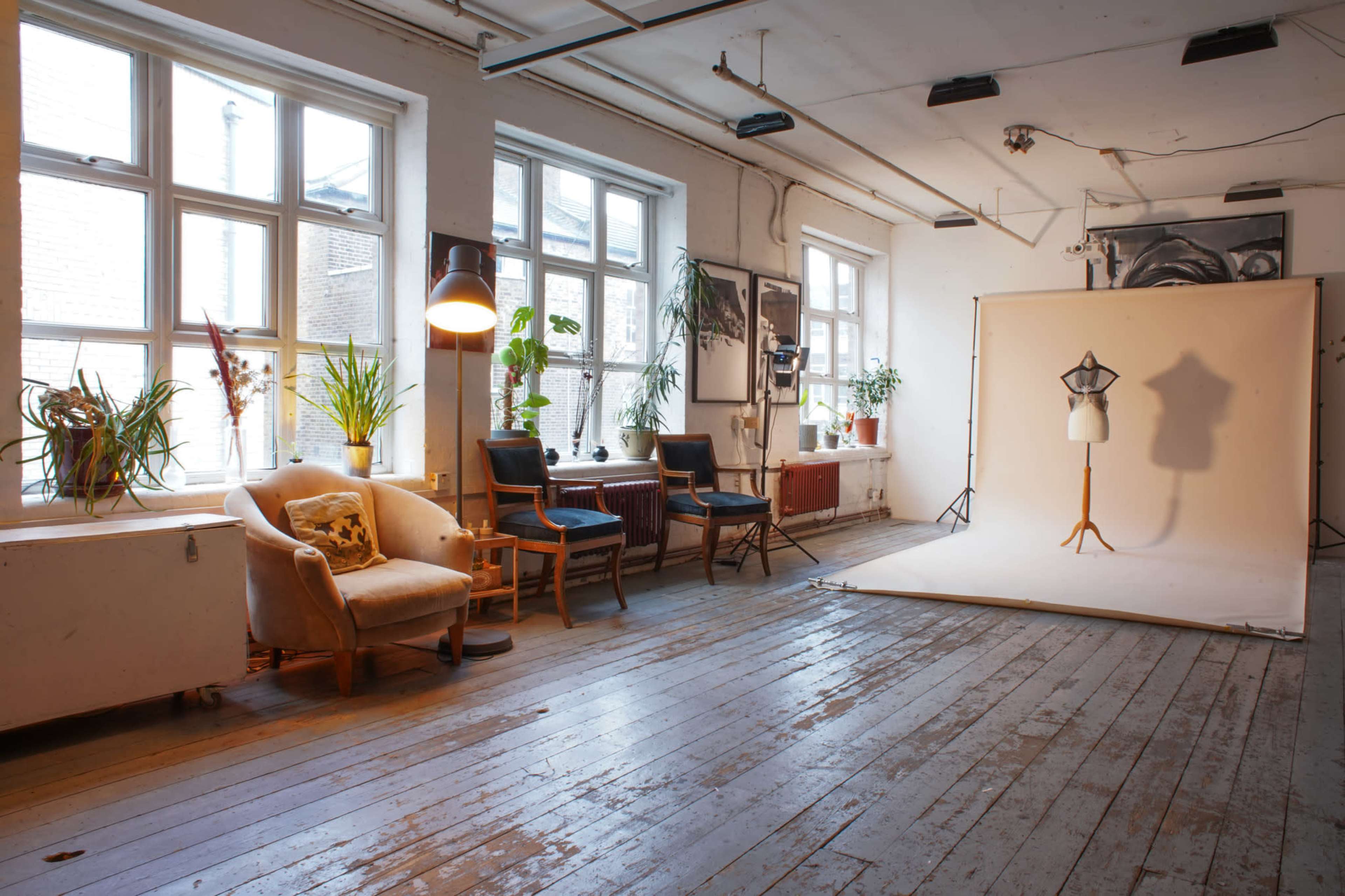 The image shows a spacious studio with large windows, featuring a seating area, plants, and a photography backdrop set up for a photoshoot.