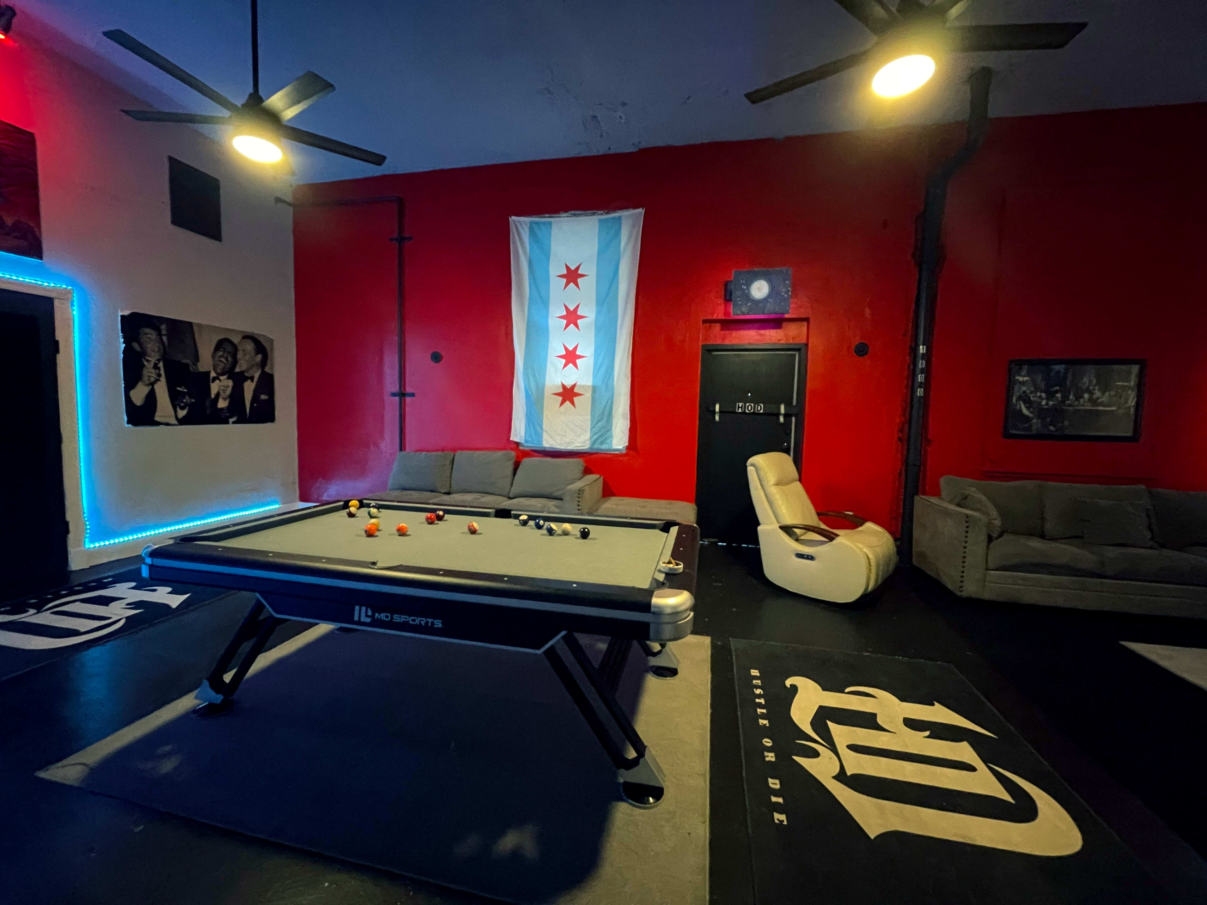 The image shows a game room with a pool table in the center, featuring a Chicago flag on the red wall and two couches in the background.