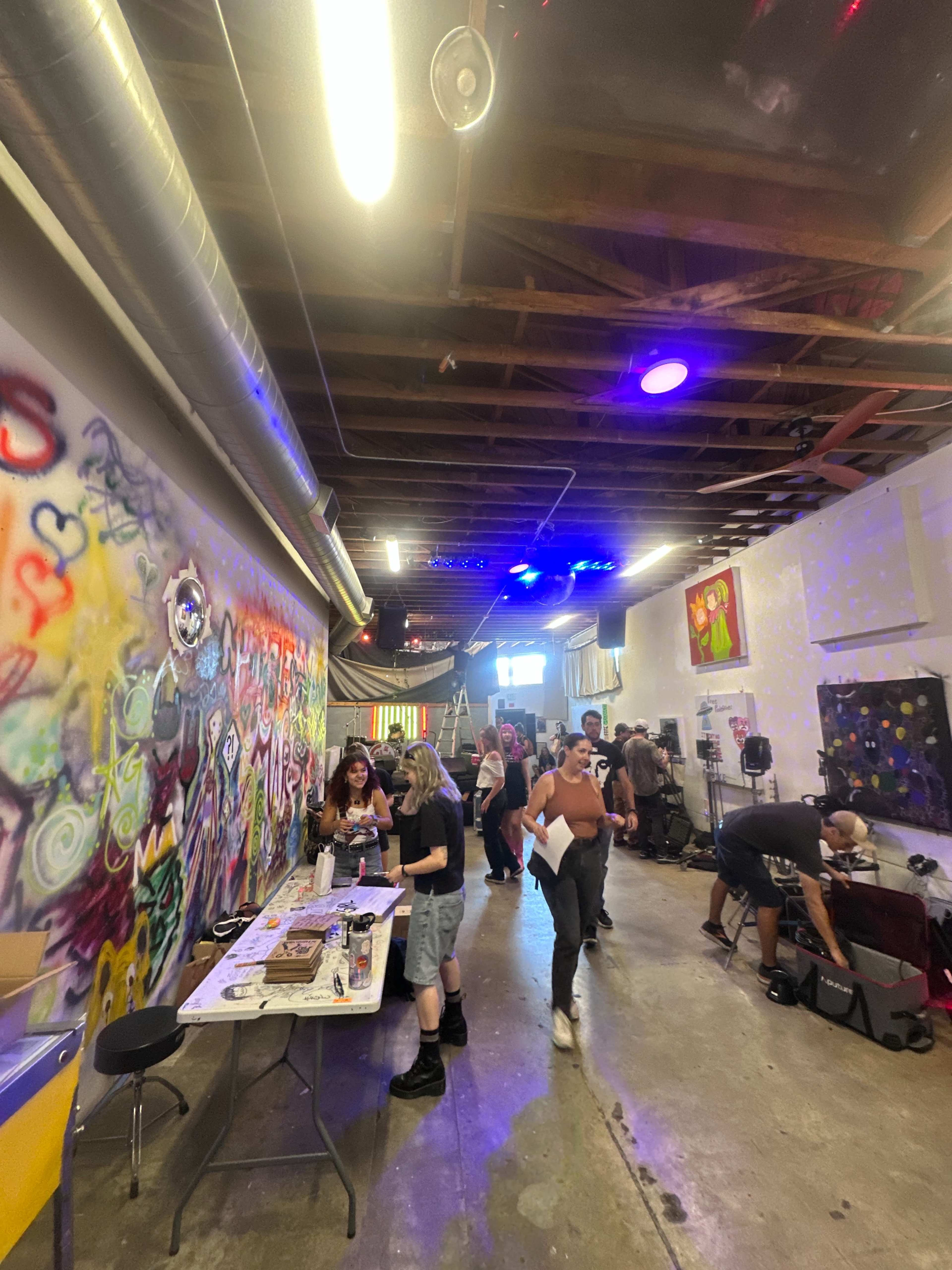 The image shows a vibrant, artsy space with people engaged in various activities amidst a colorful mural and studio equipment.