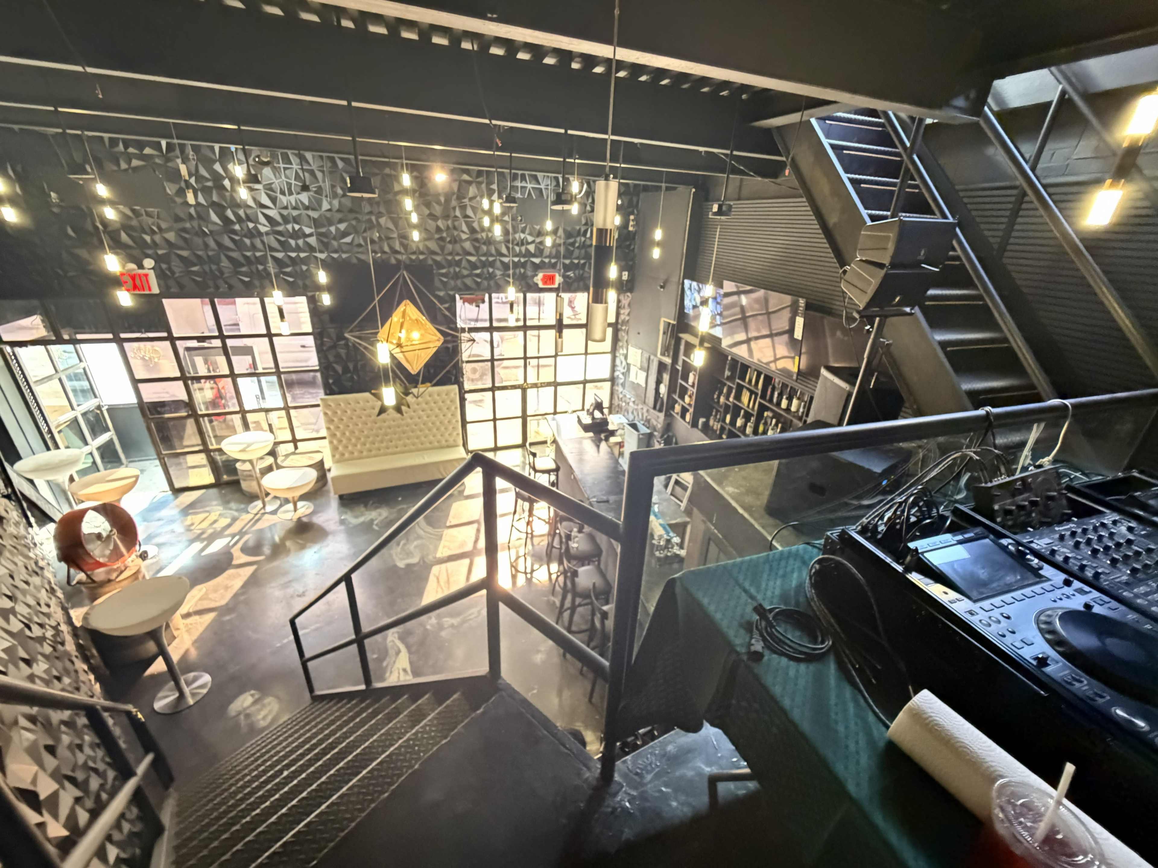 The image shows a view from an upper level of a modern venue with a bar area, seating, and spiral stairs leading down.