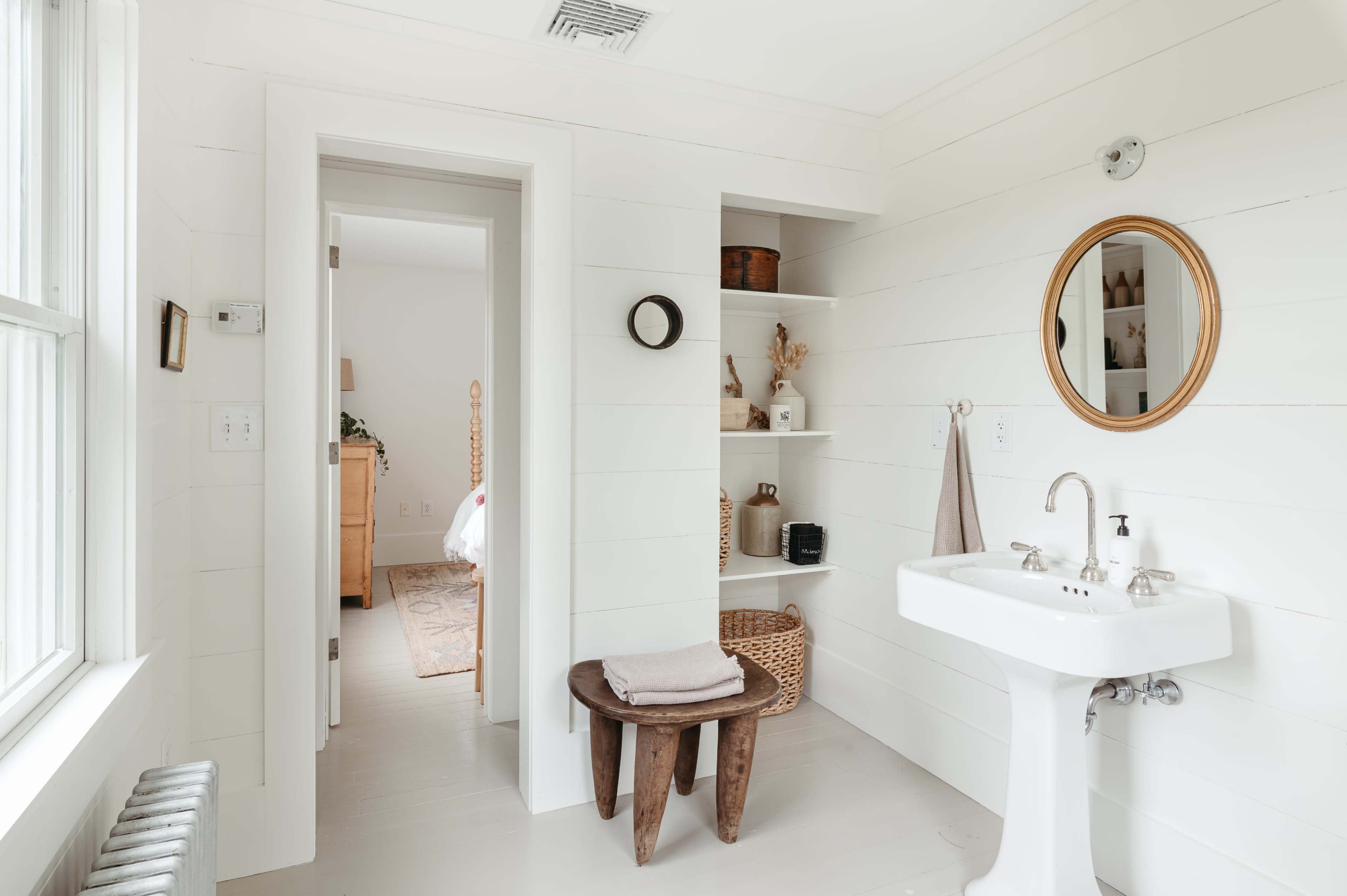 A bright bathroom featuring a white sink, a round mirror, wooden shelves with decorative items, and a doorway leading to another room.