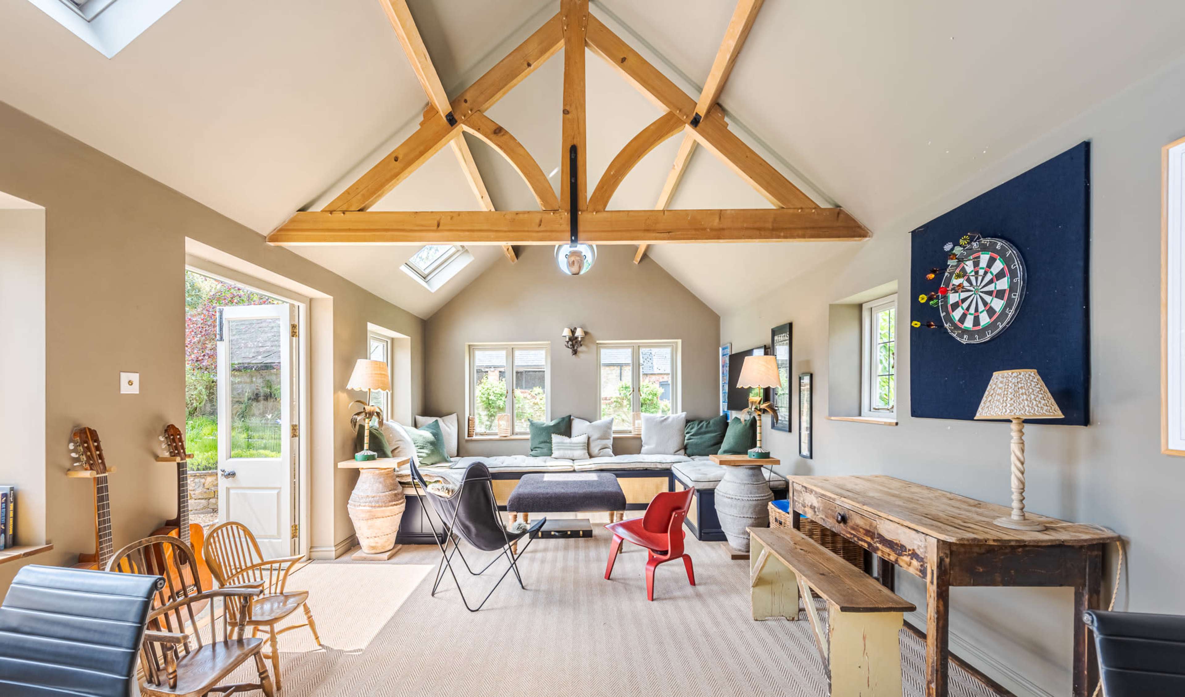 The image features a spacious living area with a high wooden beam ceiling, a large sectional sofa, dartboard, and a mix of wooden and metal furniture.