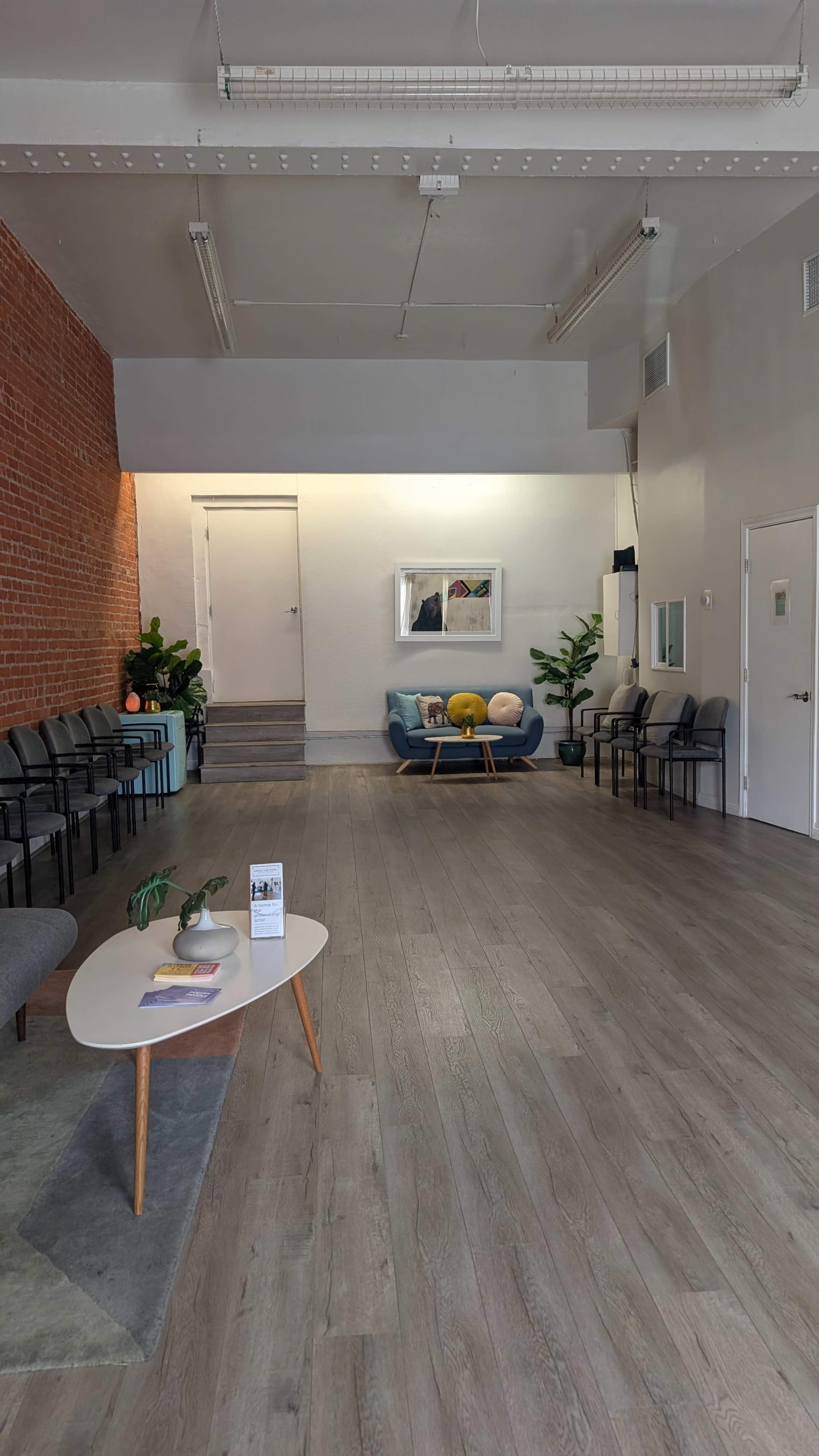 The image shows a modern waiting area with gray flooring, a couch, plants, and rows of chairs along the walls.