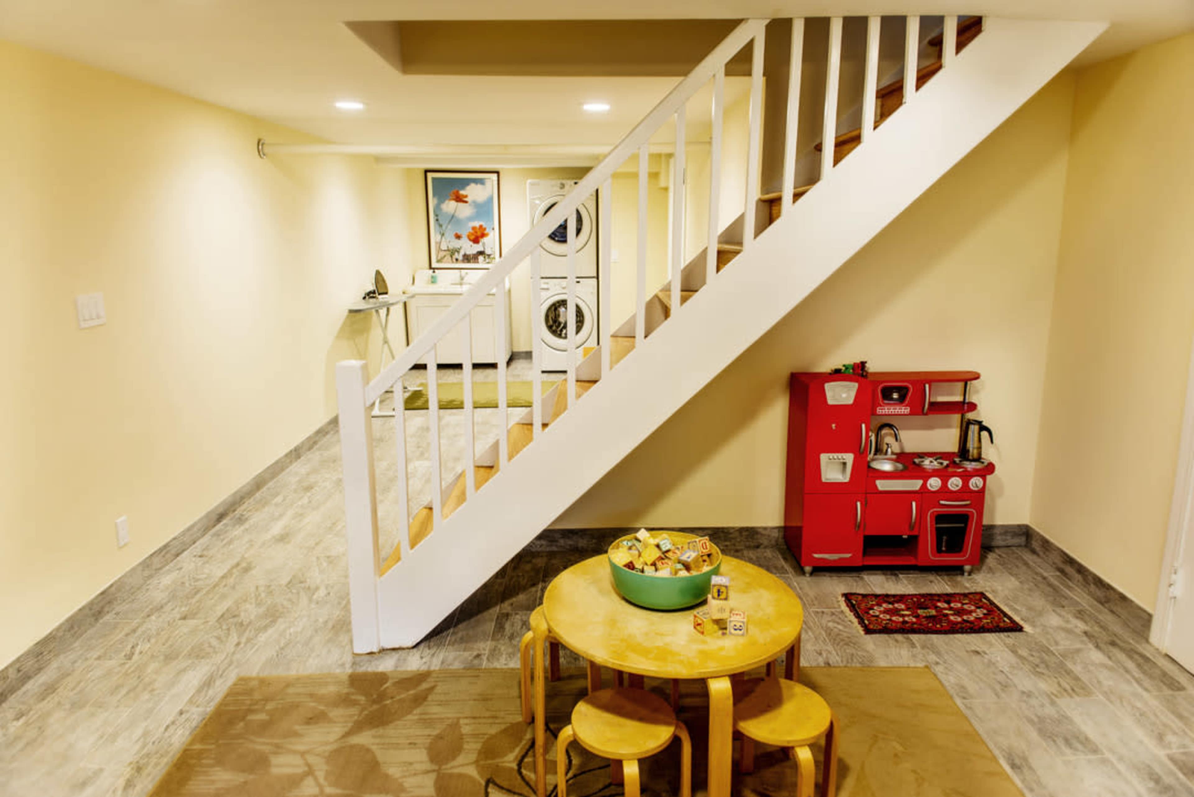 A basement area with a staircase leading to an upper level, a laundry area in the background, and a small play kitchen set along with a round table and chairs in the foreground.