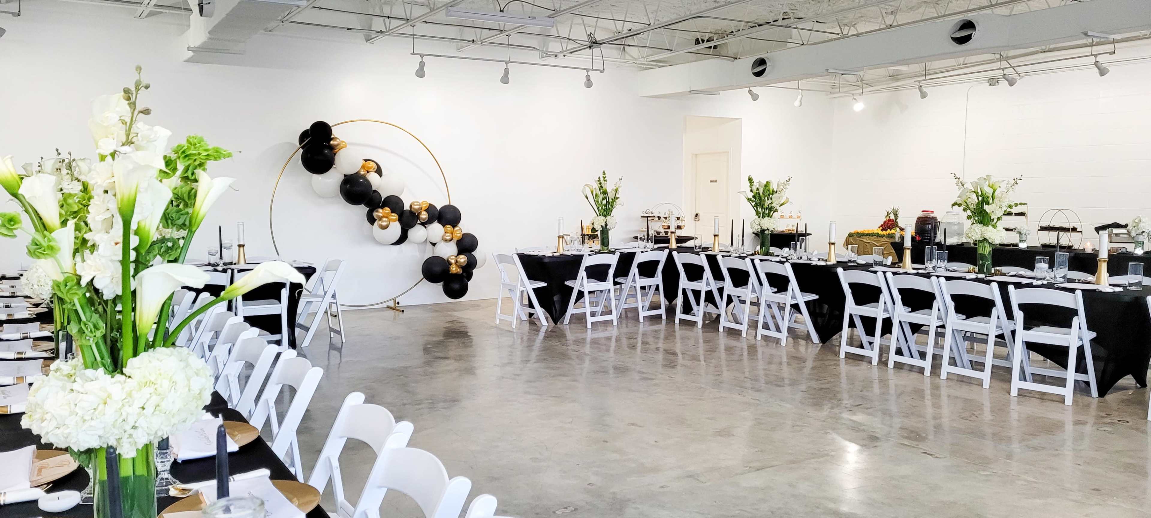 A spacious event venue set up for a formal gathering, featuring long tables adorned with black tablecloths, white chairs, and decorative floral arrangements.