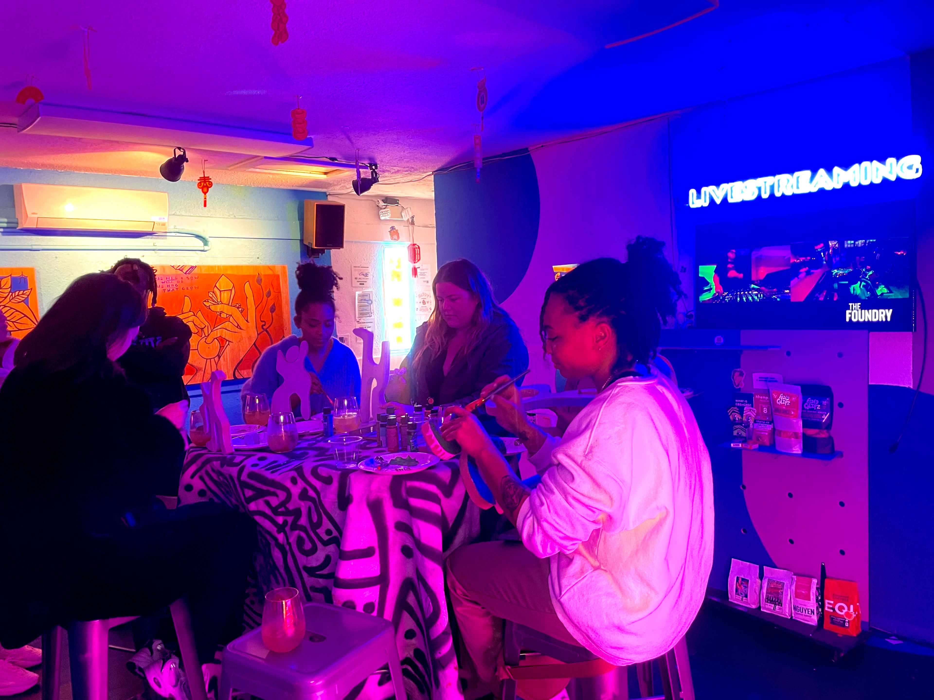 A group of people sits around a table in a colorful room, engaged in conversation, while a screen displays “LIVESTREAMING.”