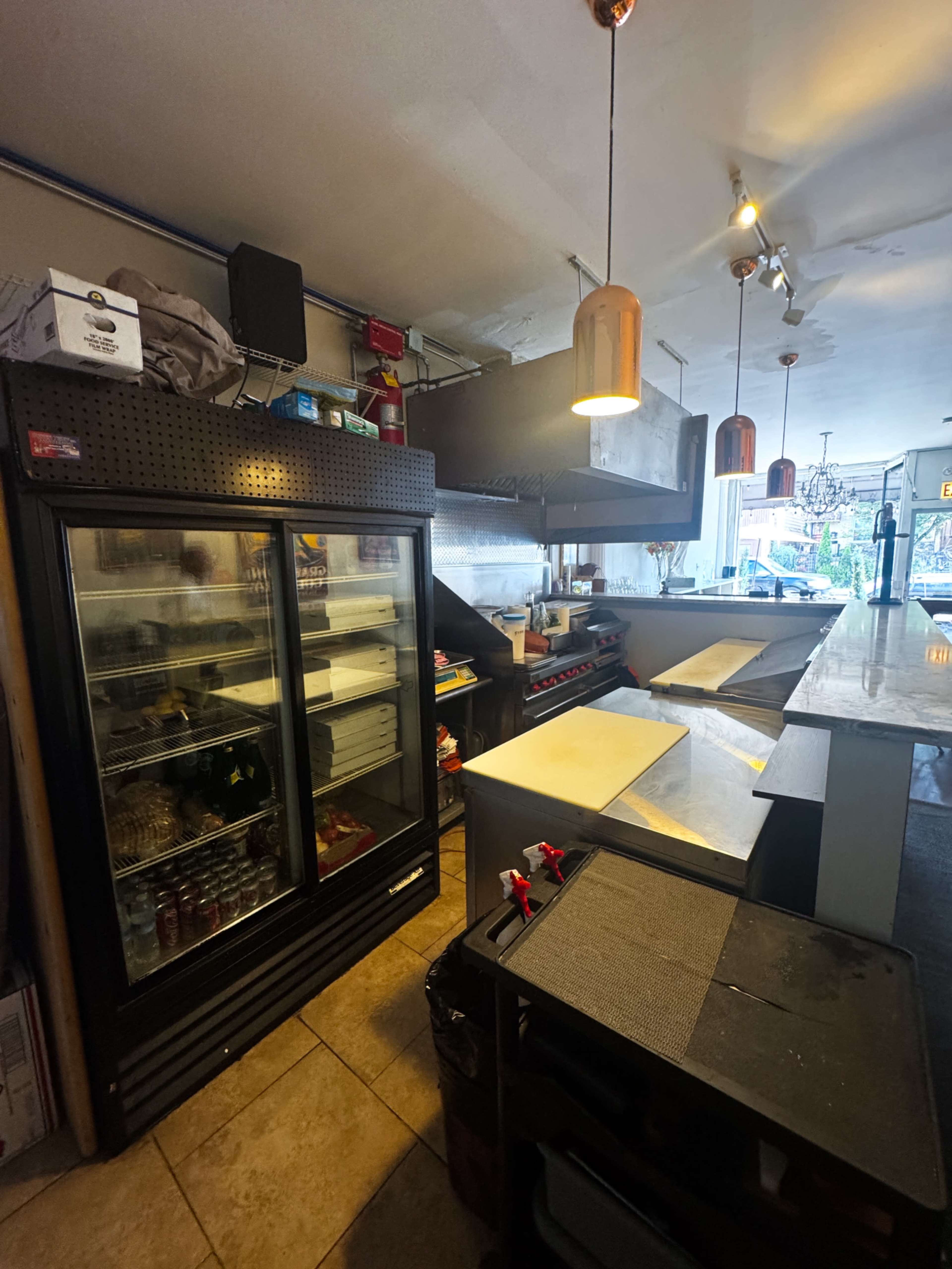 The image shows the interior of a commercial kitchen with a glass-front refrigerator, stainless steel countertops, and overhead lights.