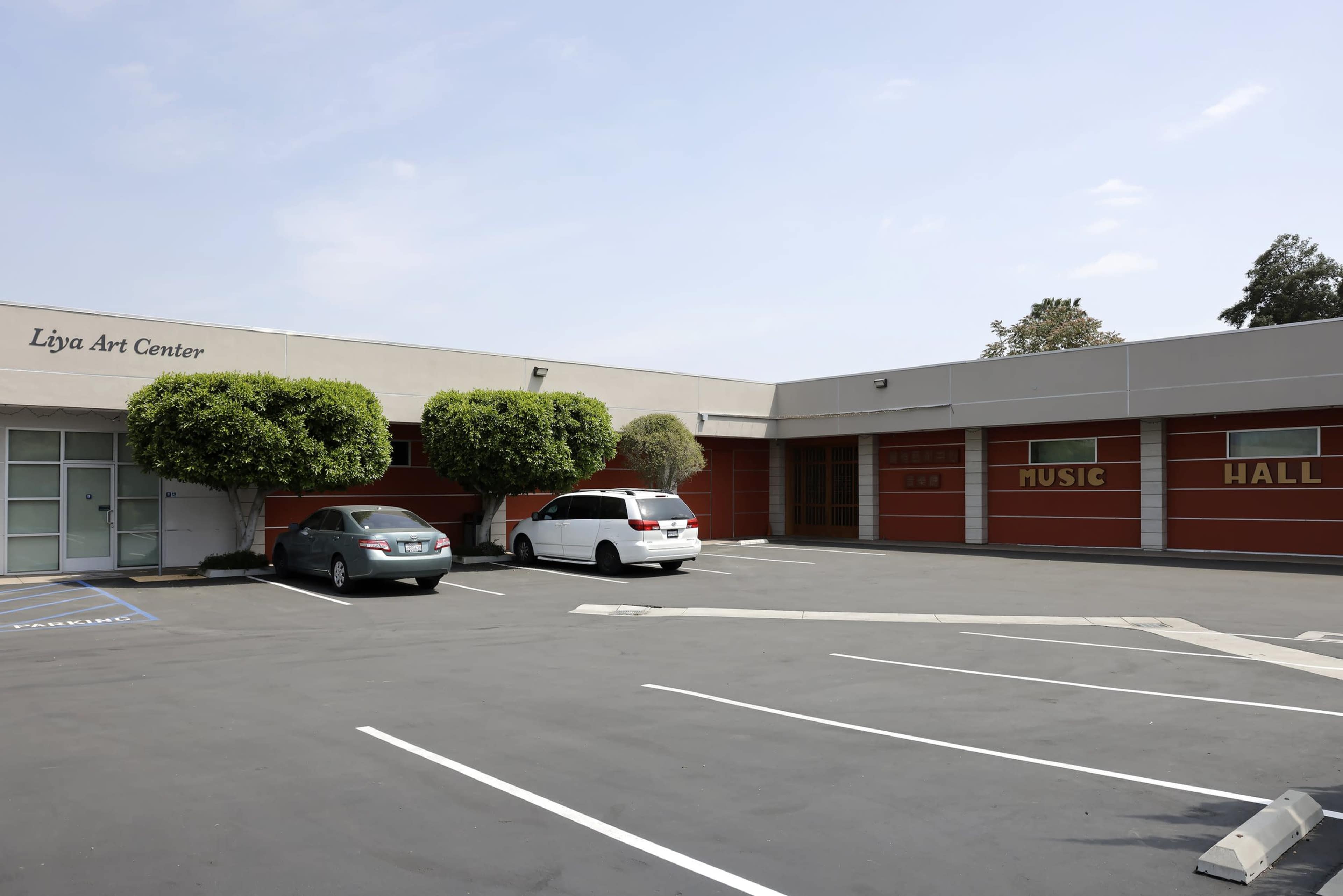 The exterior of the Liya Art Center, featuring a parking lot with several parked cars and a building labeled "MUSIC HALL" on one side.