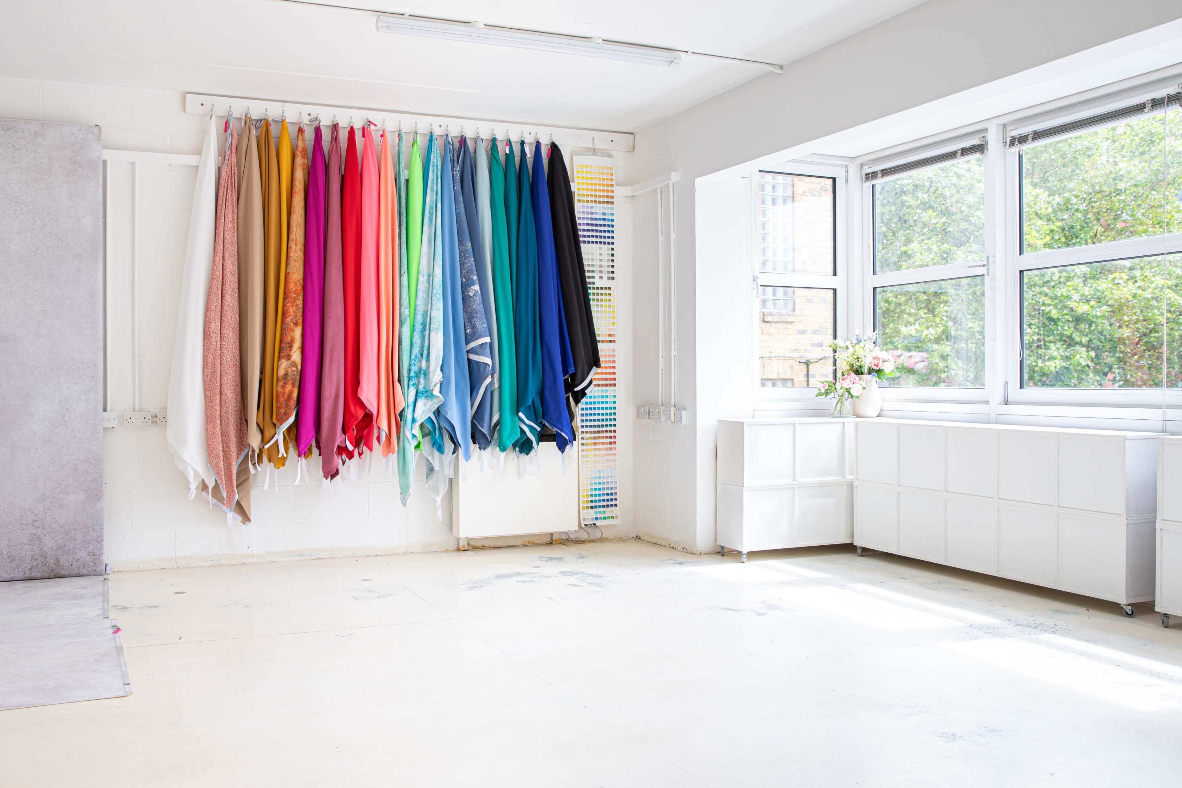 The room features a collection of colorful fabric swatches hanging on a rail against a white wall, with large windows allowing natural light to illuminate the space.