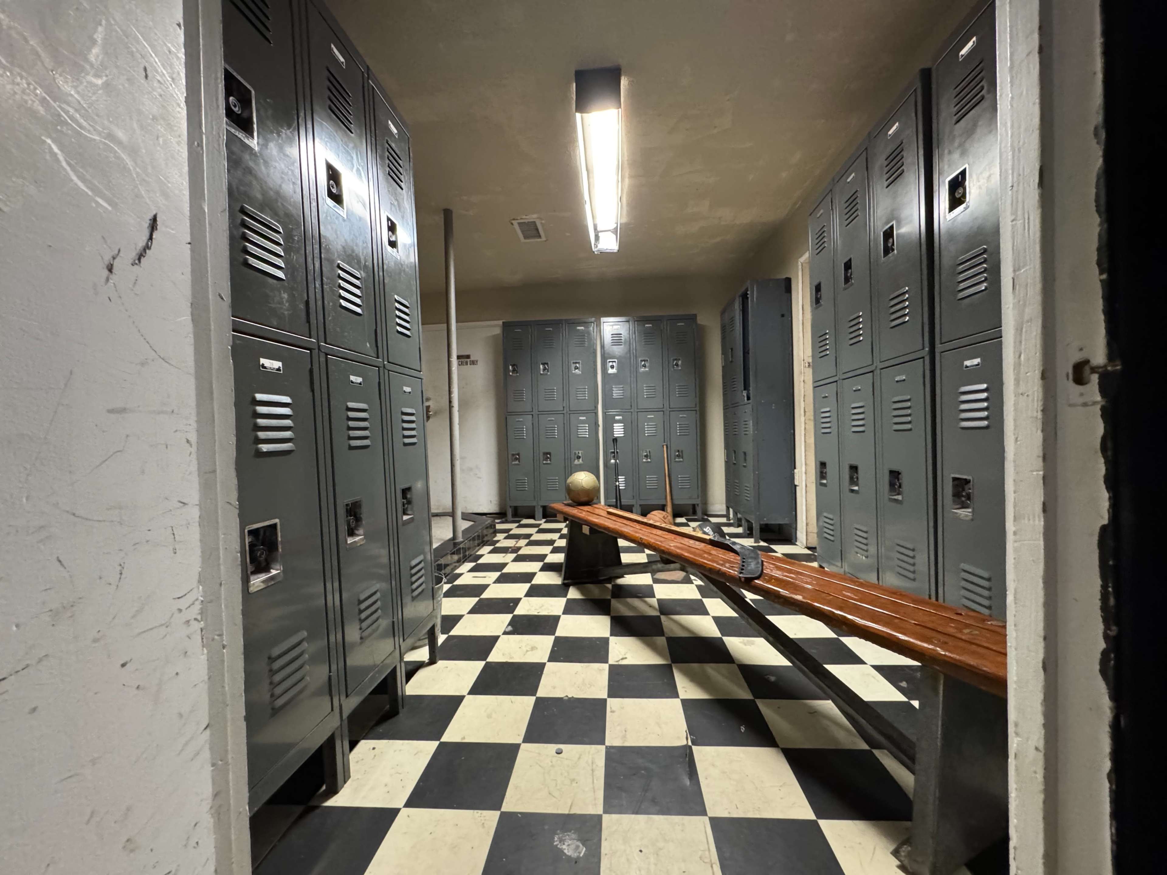 The image shows a dimly lit locker room with gray metal lockers on both sides and a wooden bench in the center, set on a black and white checkered floor.