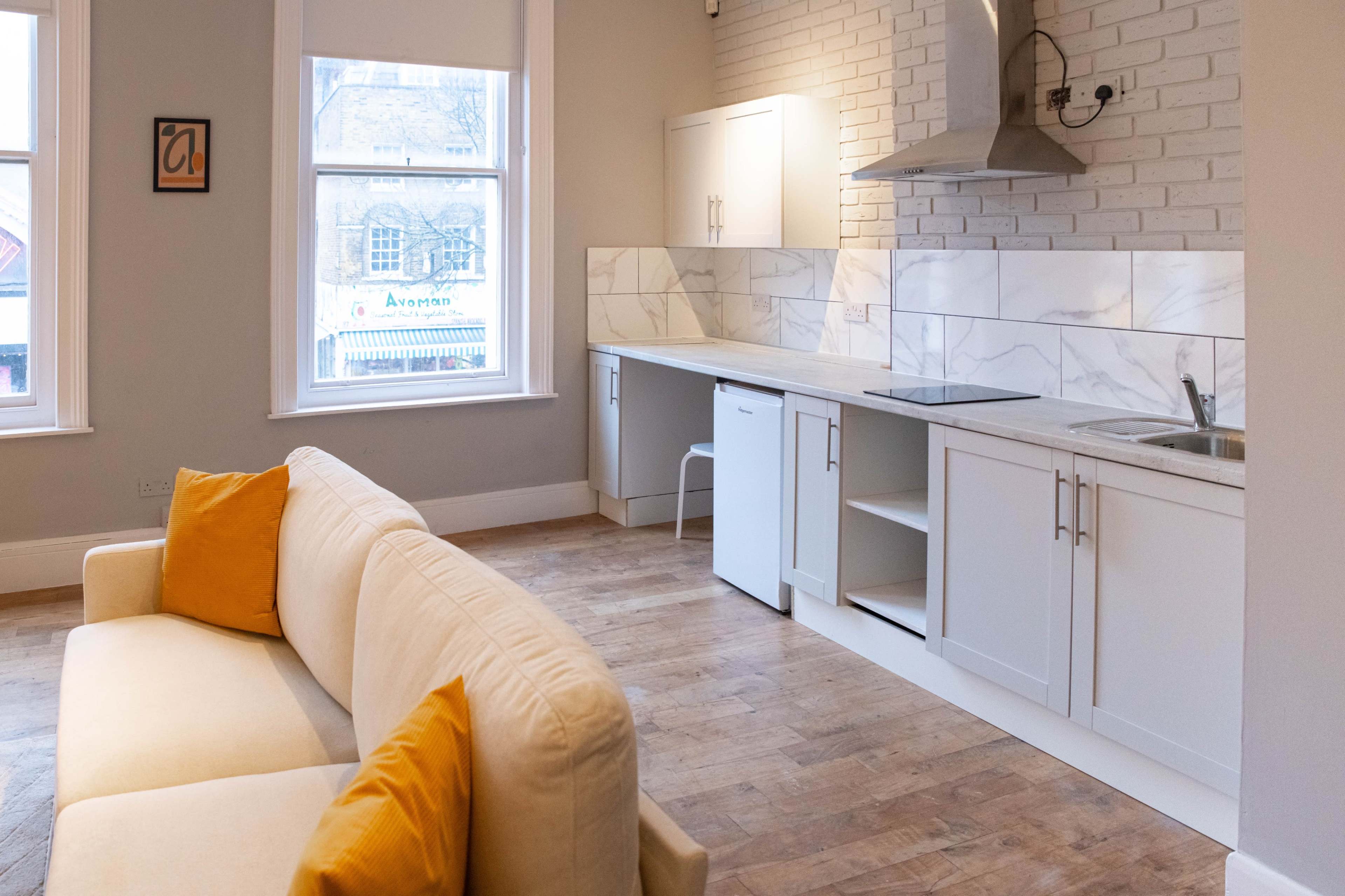 The image shows a modern kitchen and living area with white cabinetry, a marble backsplash, a stainless steel hood, and a light-colored sofa with yellow cushions.
