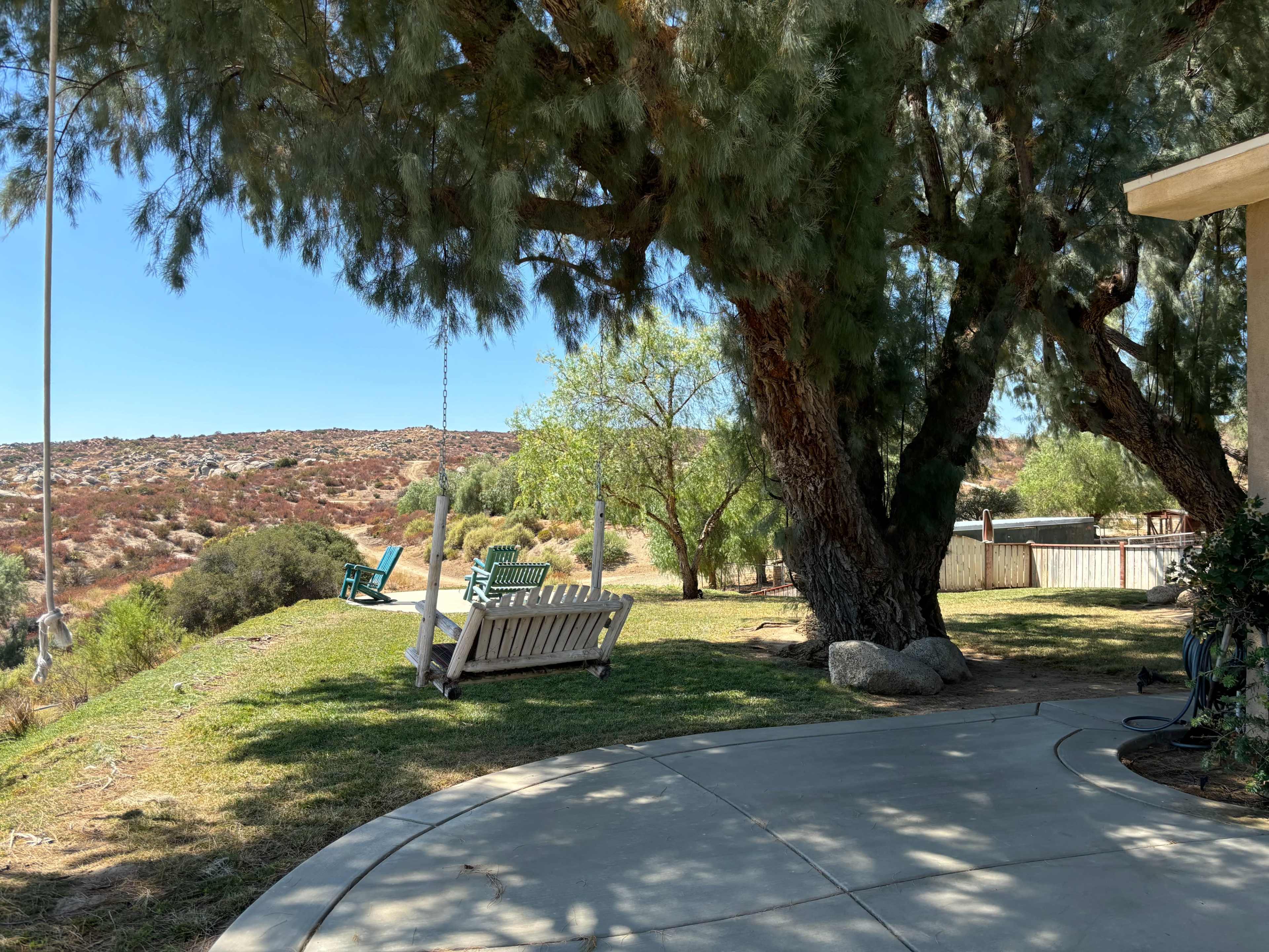 The scene features a grassy area with a concrete patio under large trees, two wooden swings, and a hillside in the background.