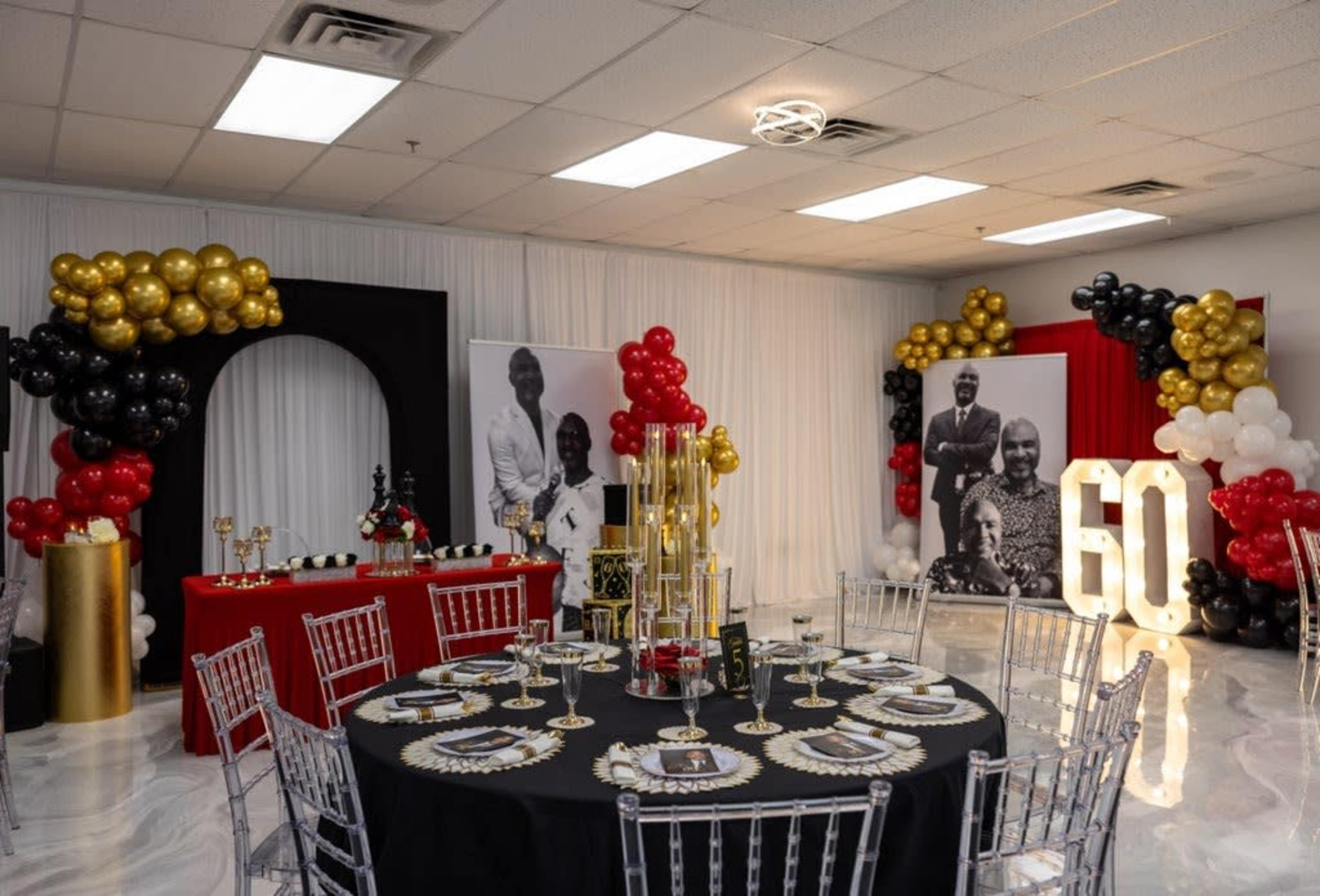 The image shows a decorated event space for a 60th birthday celebration, featuring black, gold, and red balloons, elegant table settings, and large number decorations.