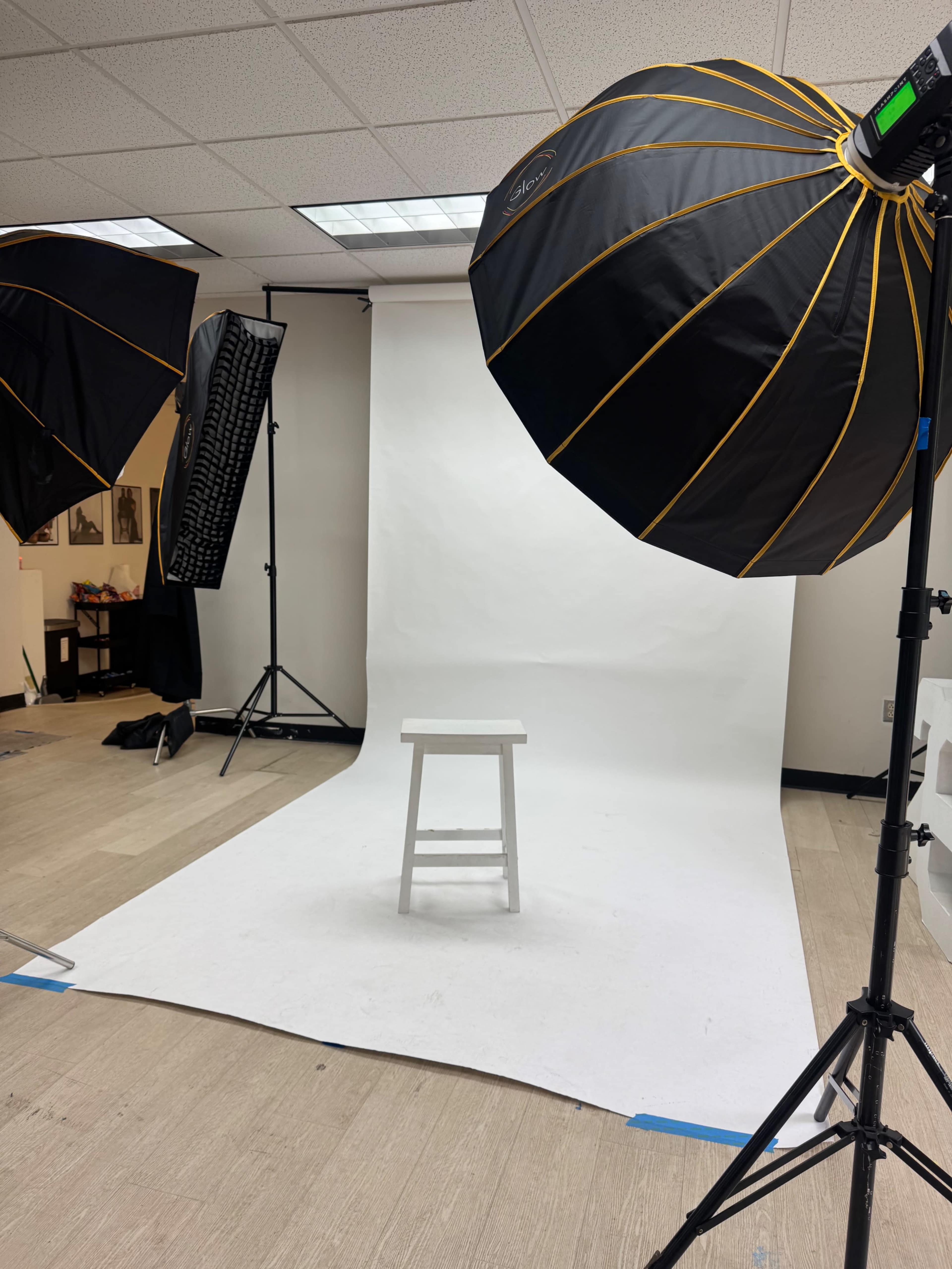 The image shows a photography studio setup with softbox lights on either side, a white backdrop, and a single wooden stool positioned in the center.
