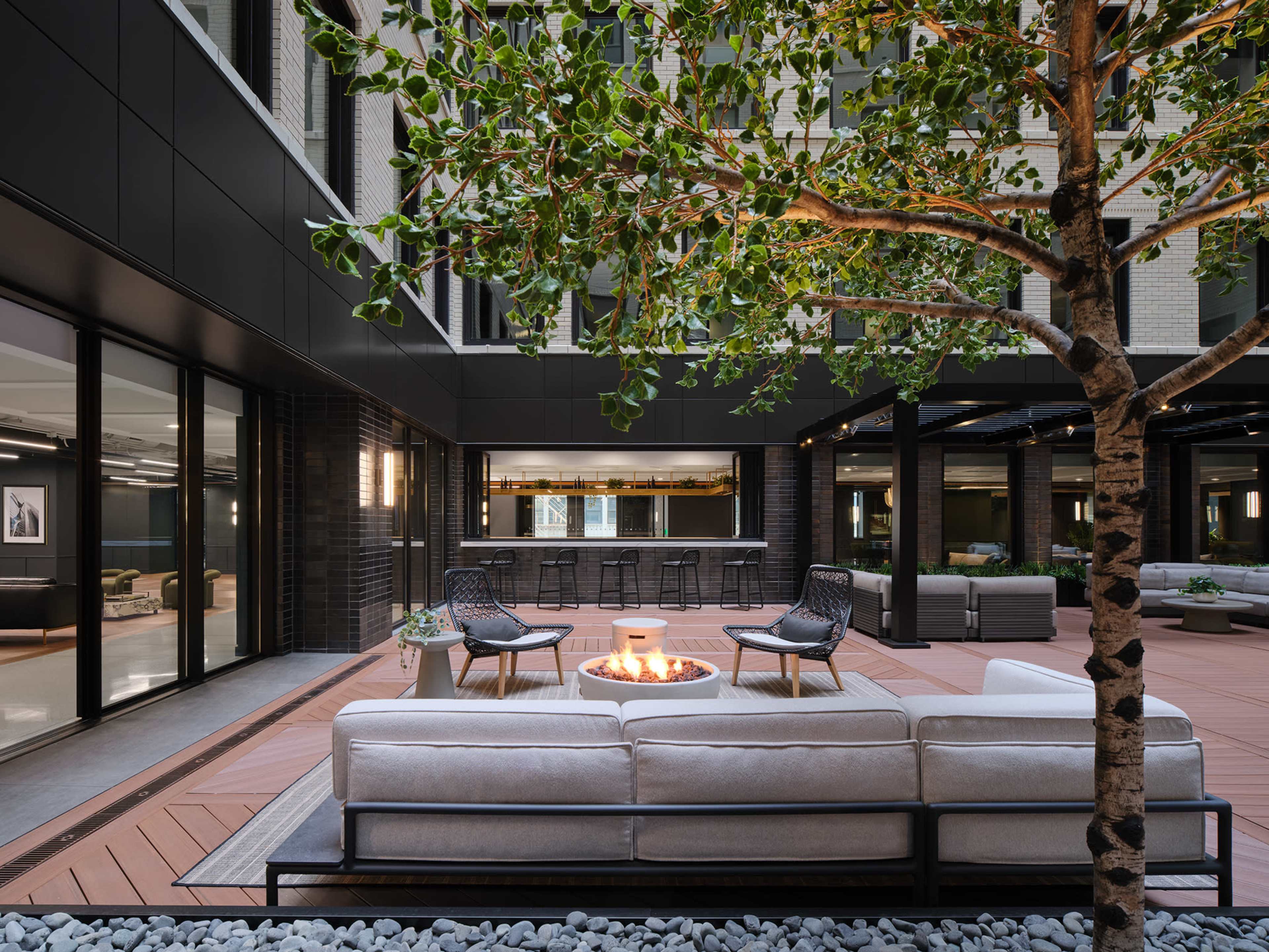 The image shows a modern outdoor lounge area with a fire pit, surrounded by seating and a tree, framed by building facades.