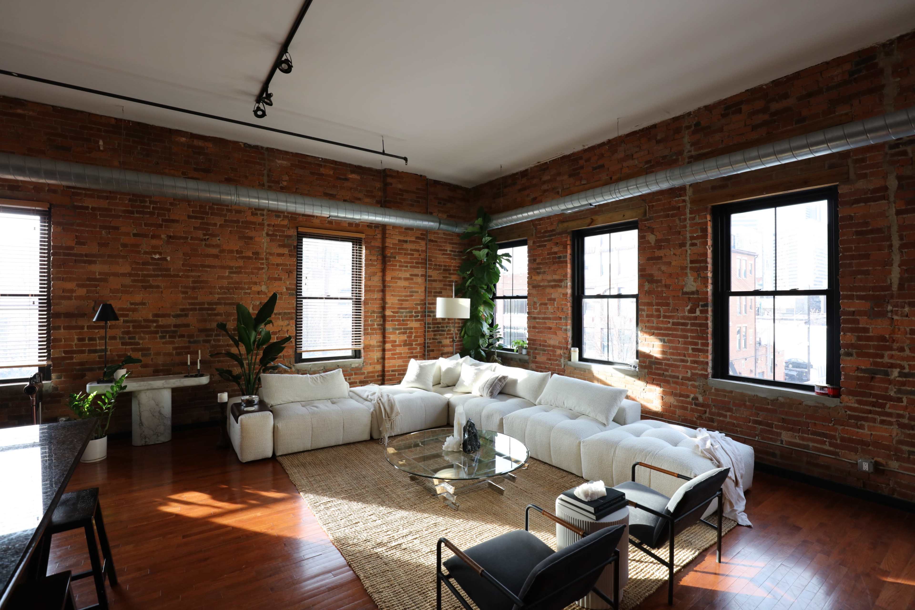 A spacious living area with exposed brick walls, large windows, a sectional sofa, and a round glass coffee table surrounded by plants and minimalist furniture.
