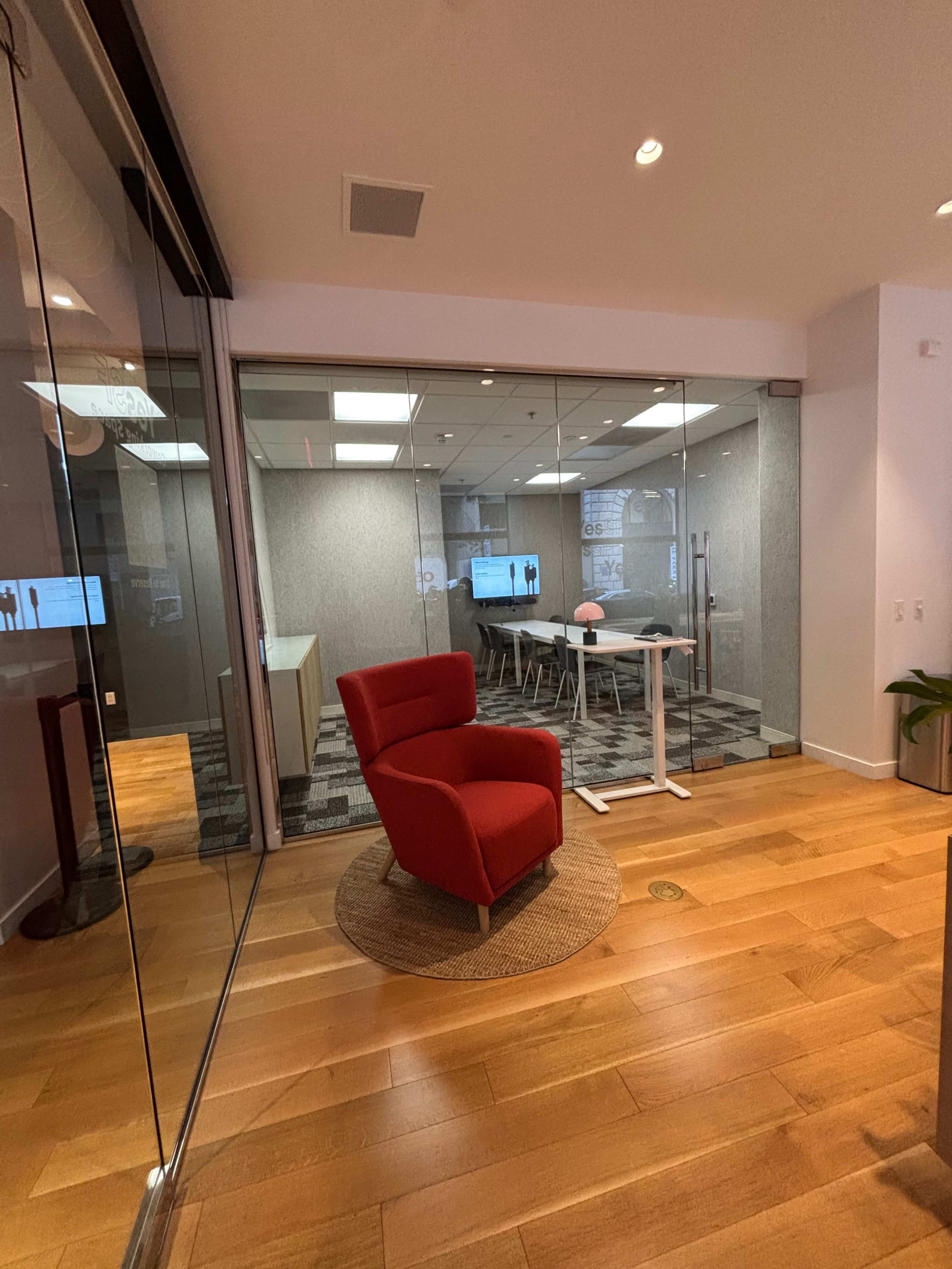 The image shows a bright office space featuring a red chair in the foreground and a meeting room with a large table and chairs visible behind glass doors.