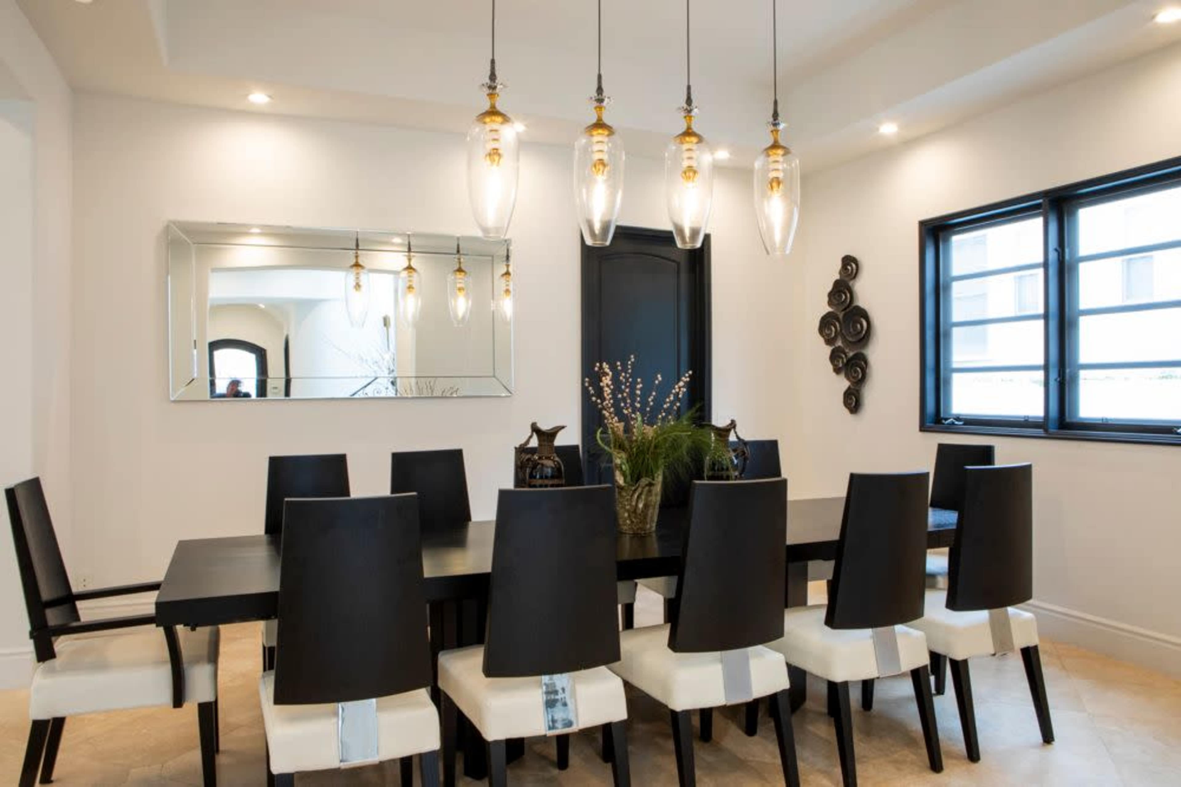 A dining room features a long black table surrounded by ten black chairs with white cushions, illuminated by pendant lights and a large mirror.