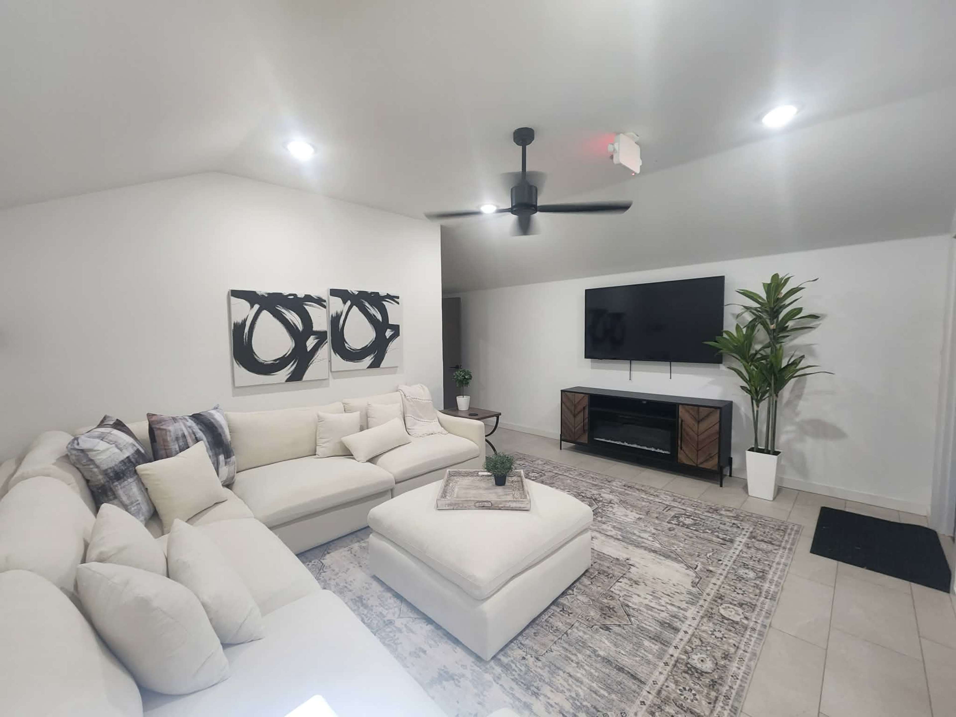 The living room features a large white sectional sofa, a matching ottoman, a modern television mounted on the wall, and a decorative plant in the corner.