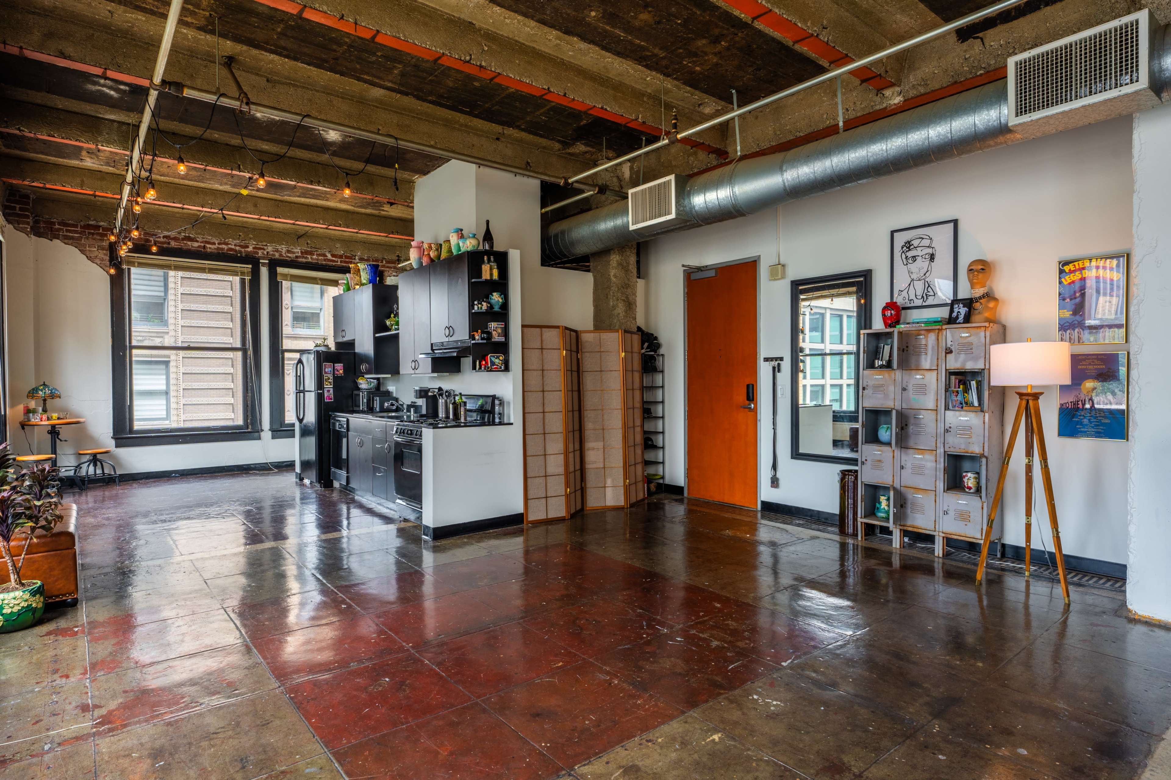 An open-concept loft with exposed brick walls, concrete ceilings, and a modern kitchenette along one side.