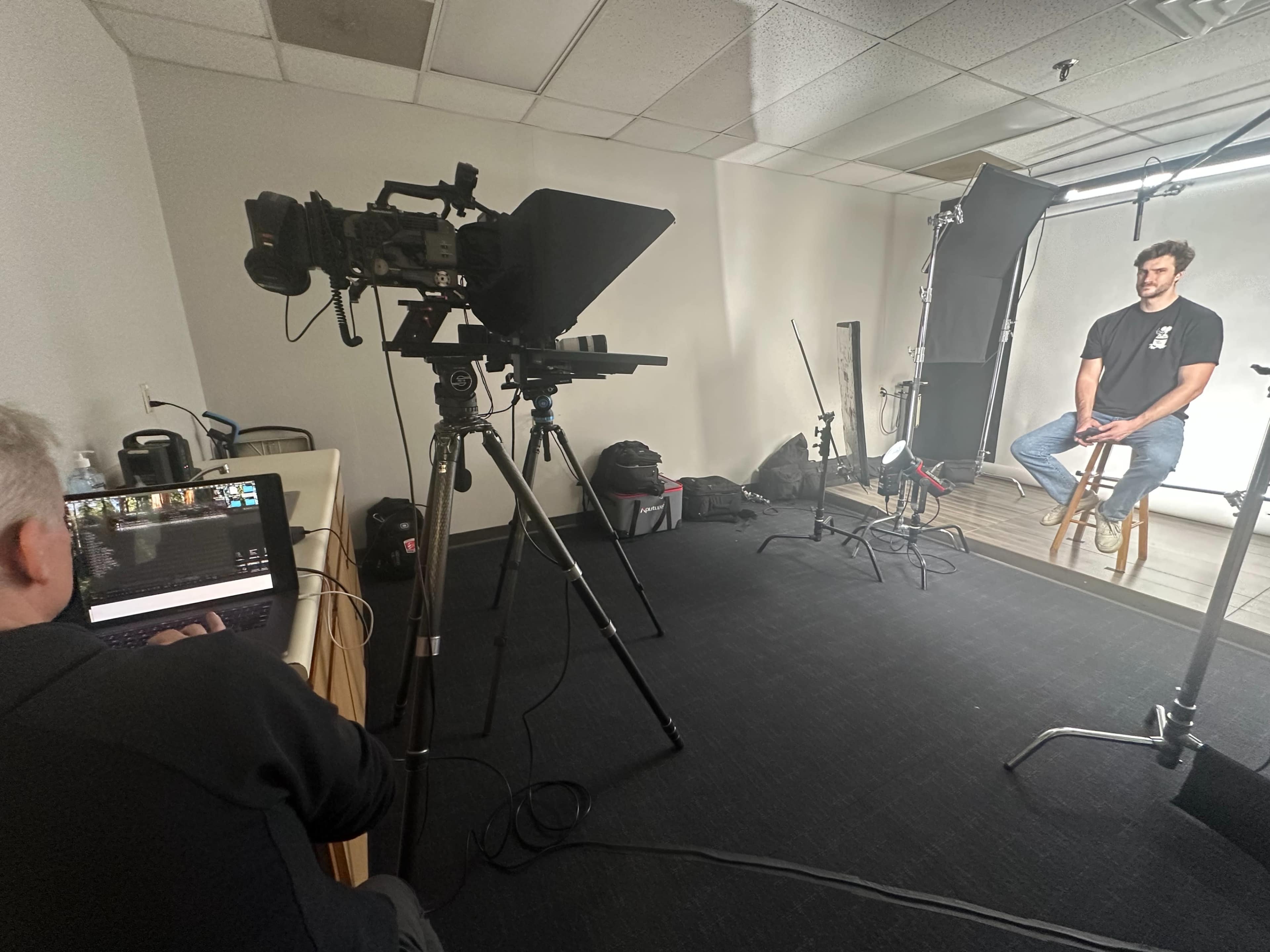 A video production setup features a camera on a tripod facing a man sitting on a stool, while a person operates a laptop in the foreground.