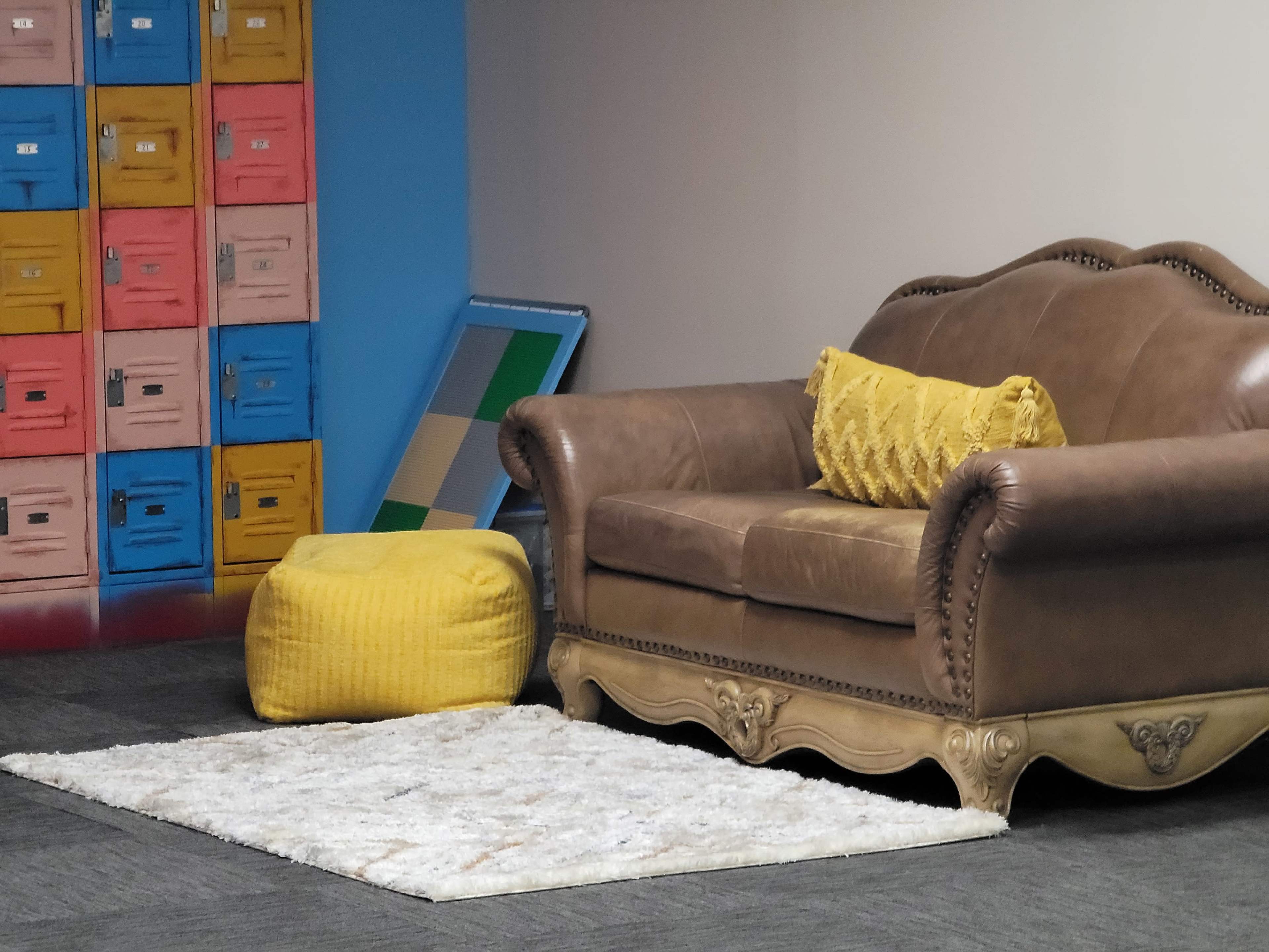 A brown leather sofa with a yellow decorative pillow, paired with a yellow pouf and a white rug, set against a wall with colorful storage lockers.