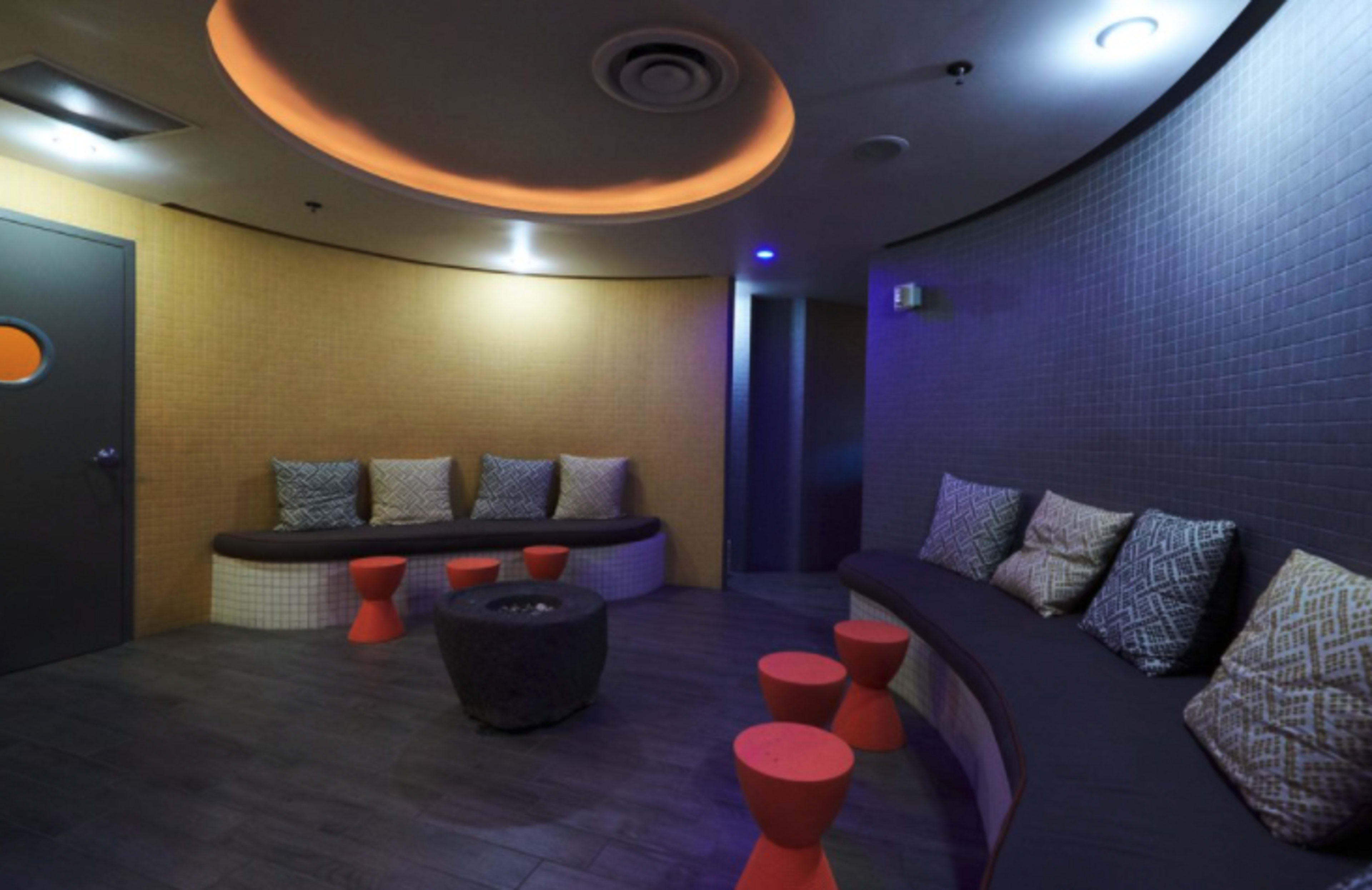The image shows a cozy lounge area with curved seating, colorful cushions, and small circular tables, illuminated by ambient lighting.