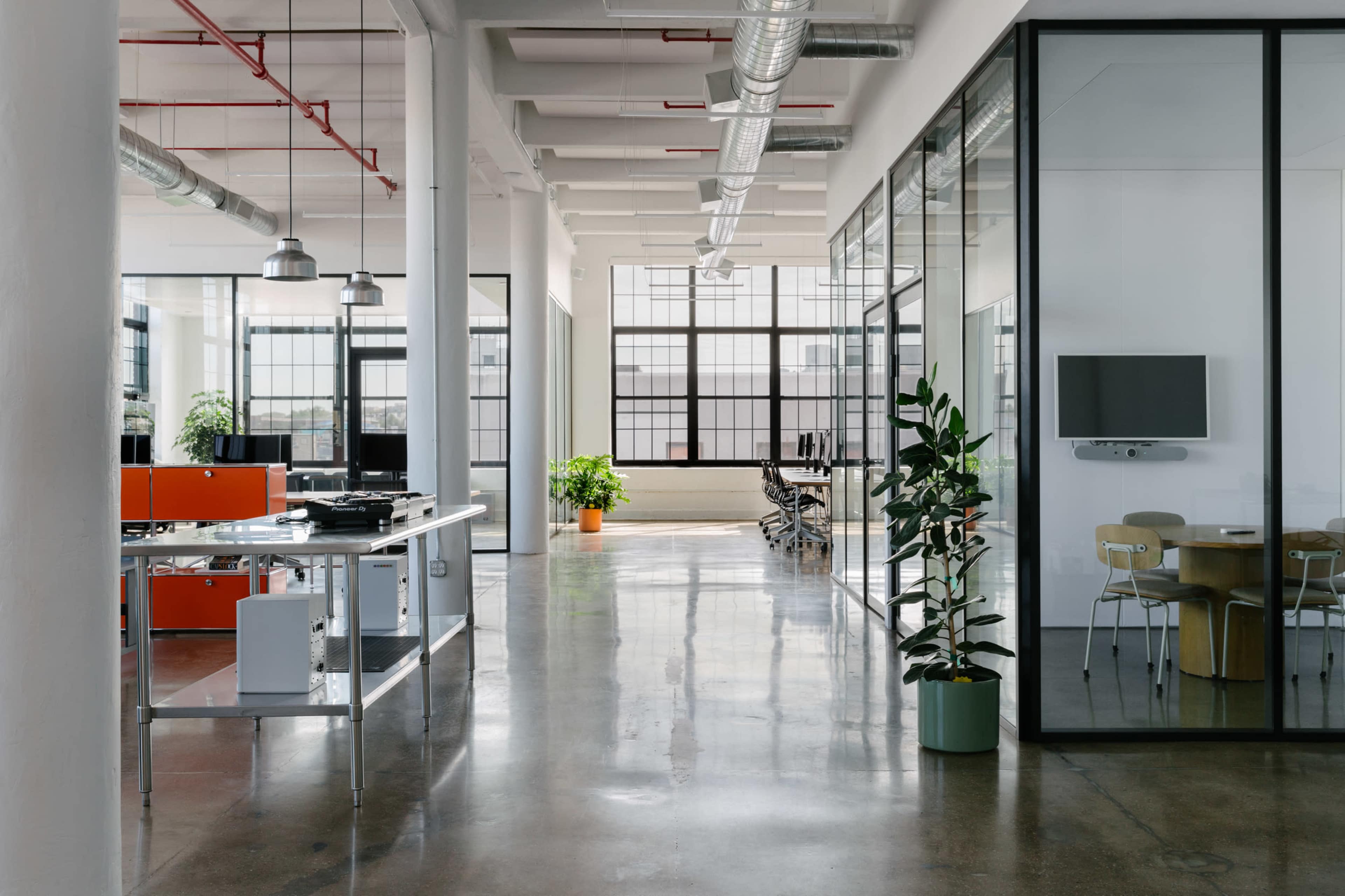 A modern office interior featuring an open layout with glass-walled meeting rooms, polished concrete floors, and plants beside workstations.